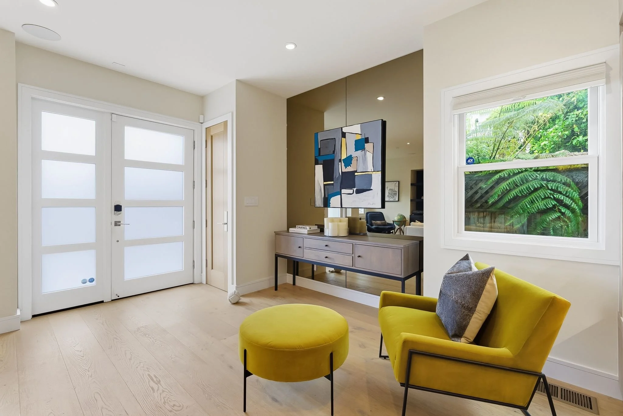 Modern entryway with a white front door with frosted glass panes, a light-colored hardwood floor, a yellow armchair with a gray pillow, a matching yellow ottoman, a sideboard with decorative items, abstract art, and a window showing greenery outside.