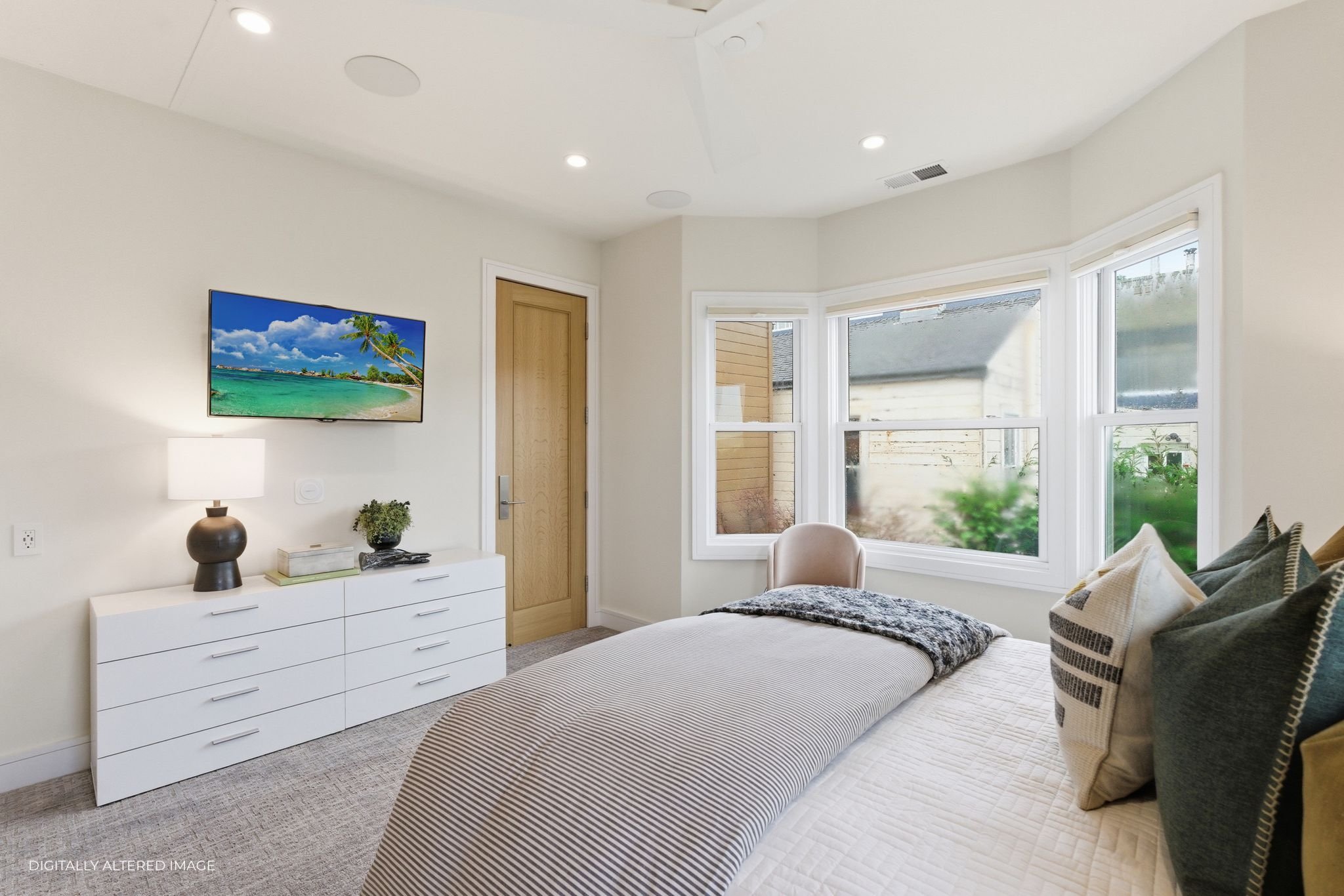 A cozy bedroom with a bed, several pillows, and a blanket. There is a white dresser with a lamp, a plant, and some books. A flat-screen TV is mounted on the wall, showing a tropical beach scene. Large windows let in natural light, and there is a wood