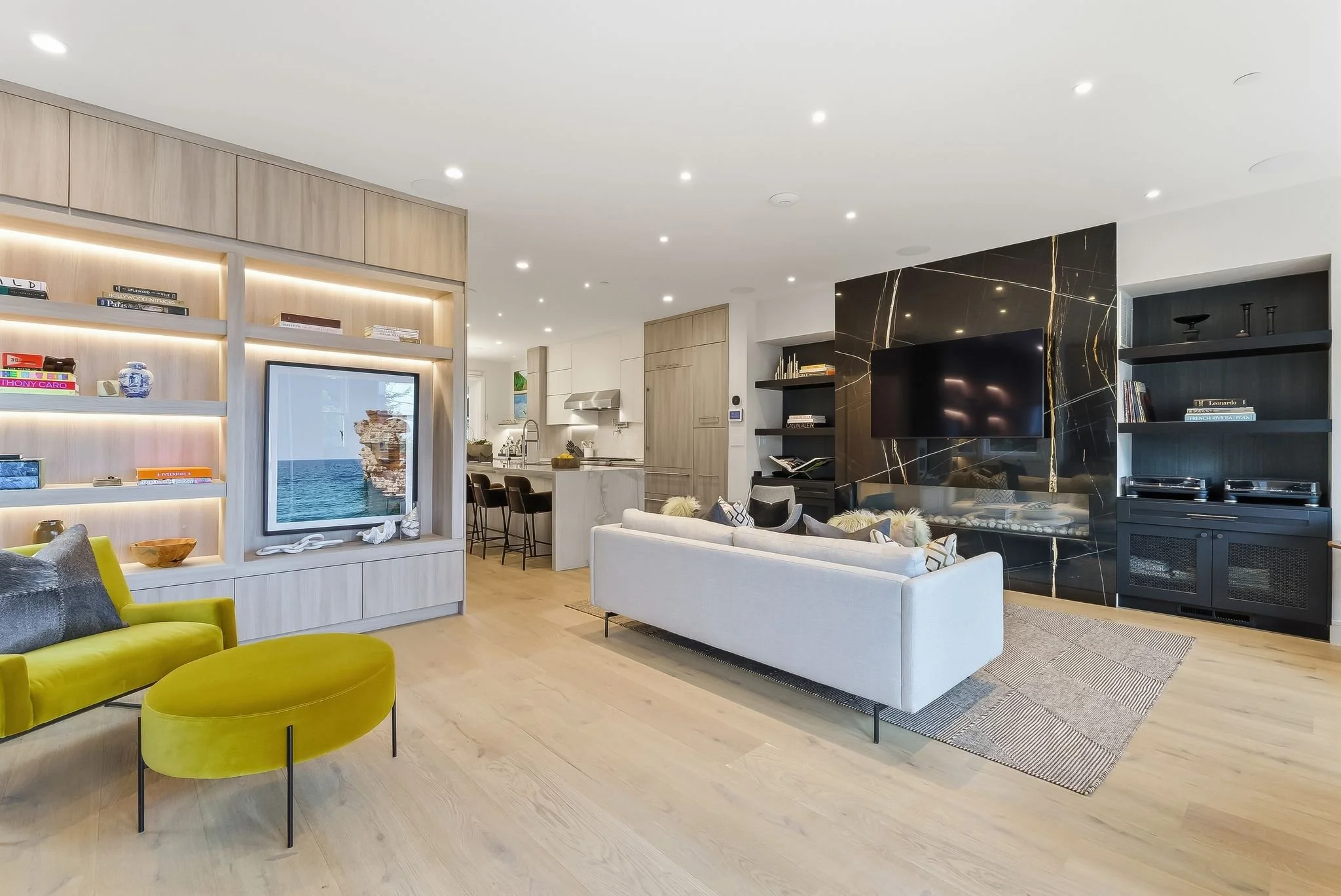 Modern living room with a white sofa, black marble accent wall with a mounted TV, yellow armchair, built-in shelves, and an open kitchen in the background.