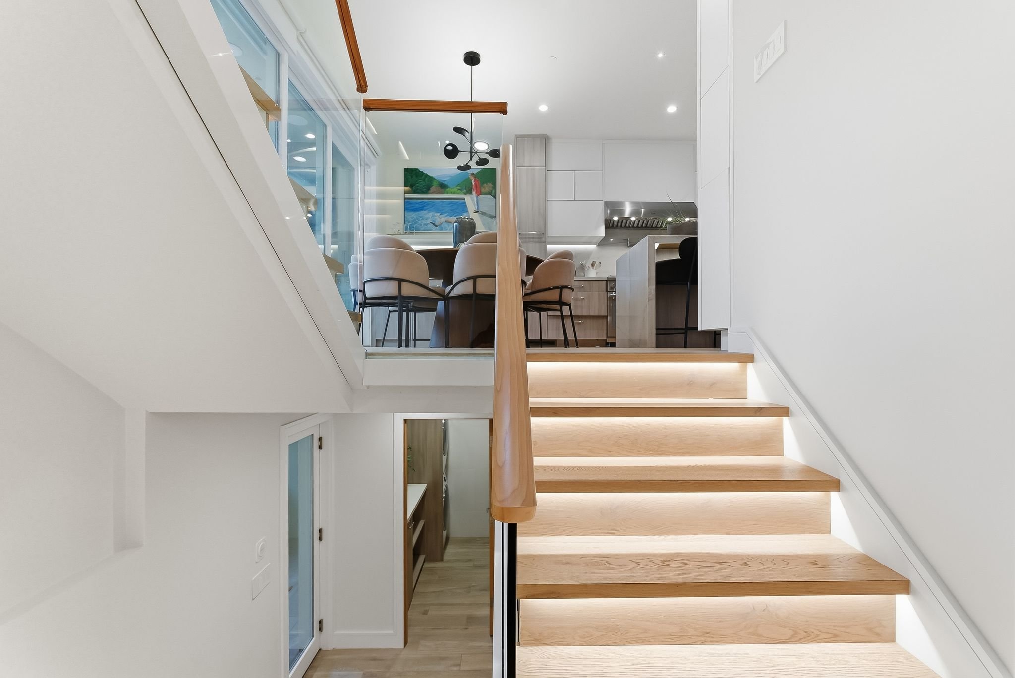 Interior of a modern home showing a staircase with wooden steps and LED lighting underneath, leading up to a dining area with beige chairs and a round wooden table, visible kitchen with white cabinets, and a view of a painting of a landscape with a p