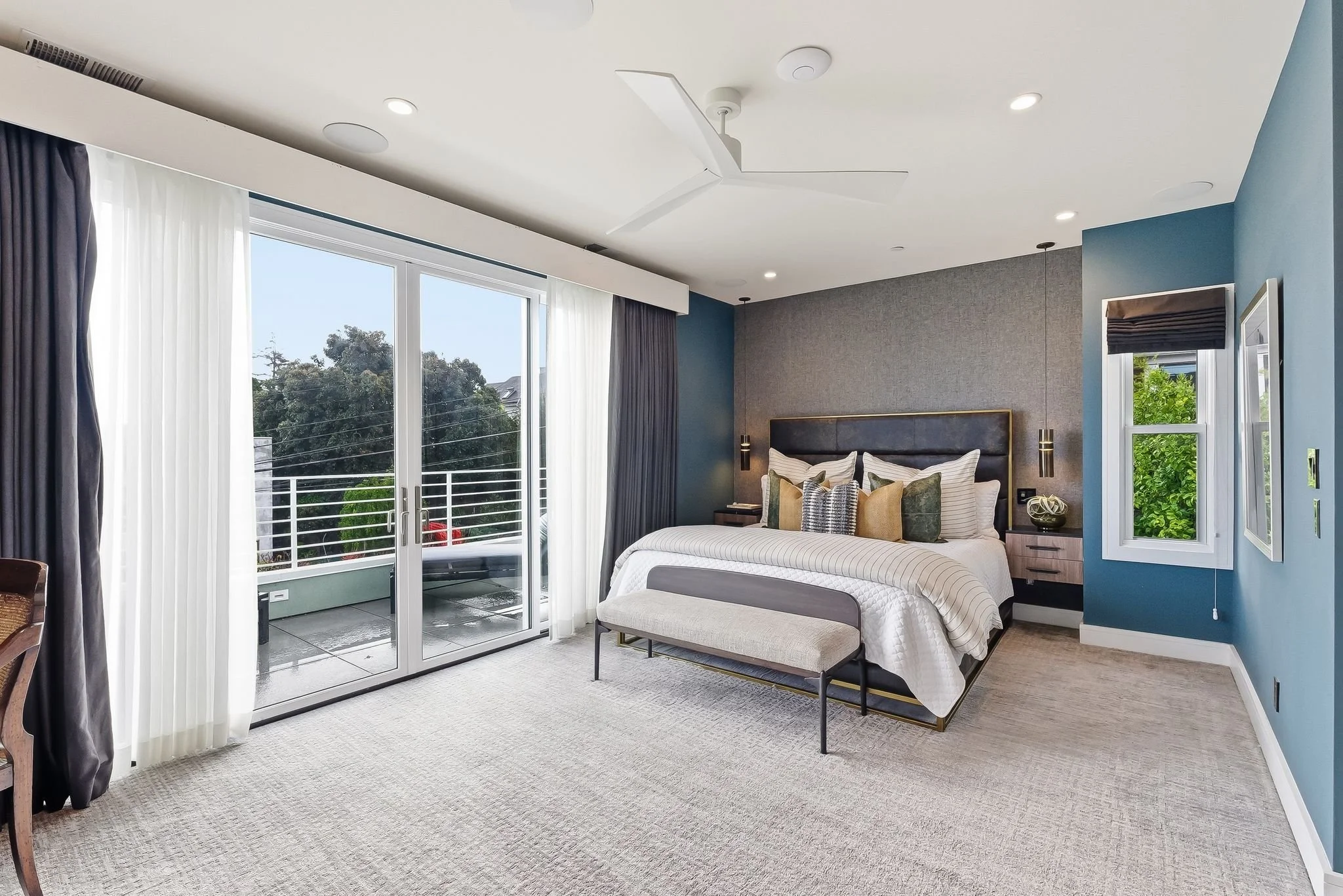 Modern bedroom with a large bed, decorative pillows, a bench at the foot of the bed, and a sliding glass door leading to a balcony with outdoor seating, trees, and a blue sky.