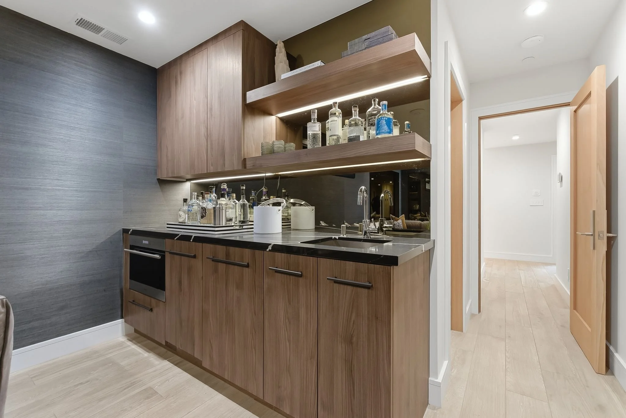 Modern kitchenette with wooden cabinets, black countertop, and open shelves with bottles of alcohol and glassware.