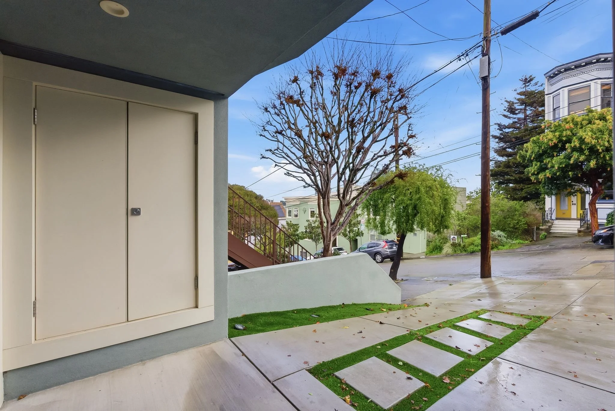 View of a residential street with a tree, parked cars, and various houses; the foreground shows a concrete sidewalk with stepping stones and small patches of grass.