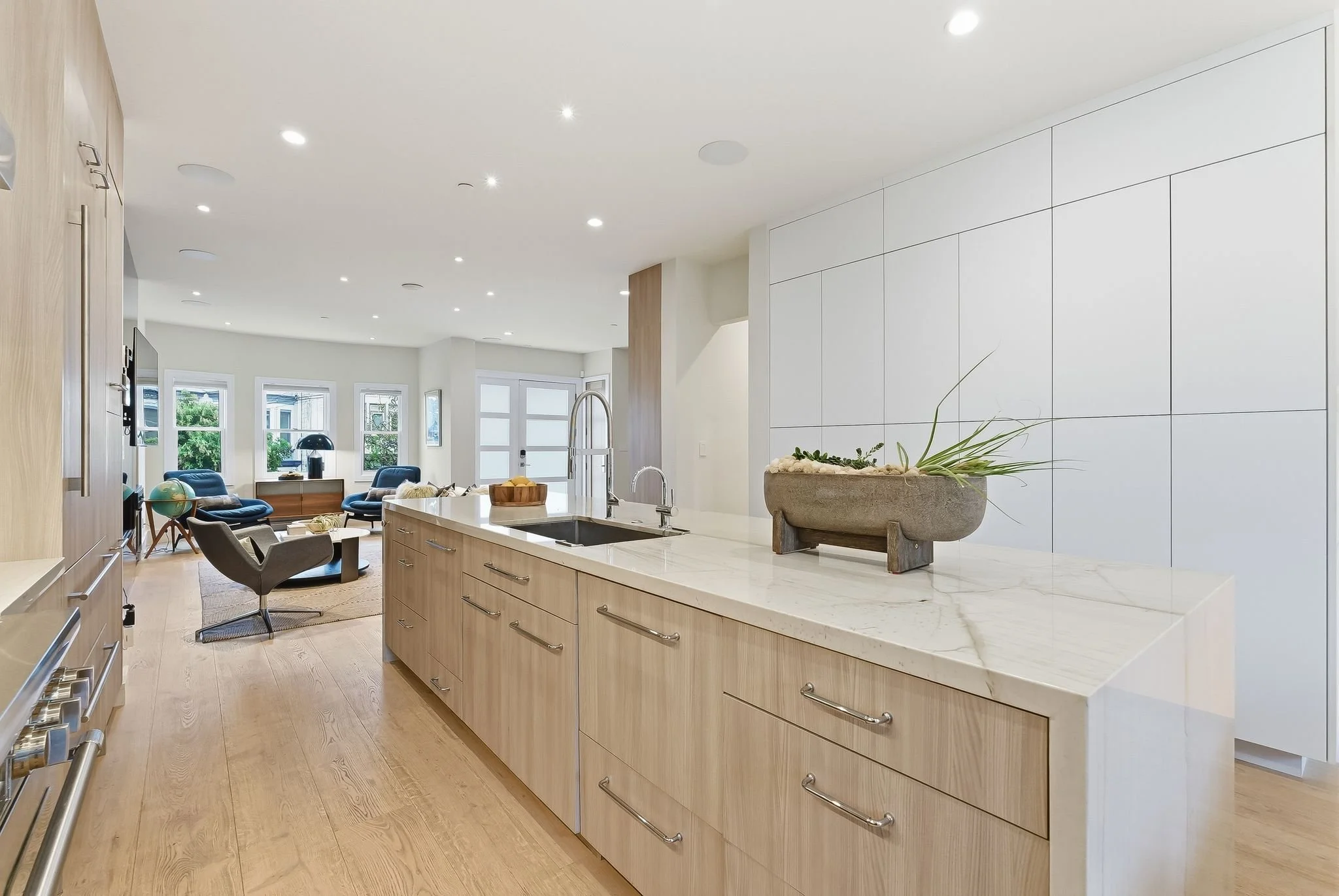 Modern open-concept kitchen with beige cabinets, white marble countertops, and a living area with blue chairs and large windows.