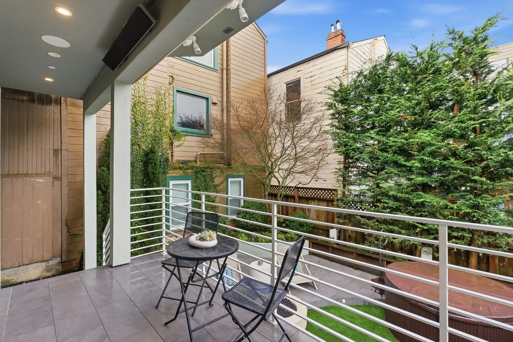 View of a small patio with black patio furniture, a white railing, and a hot tub, surrounded by trees and neighboring houses, under a blue sky.