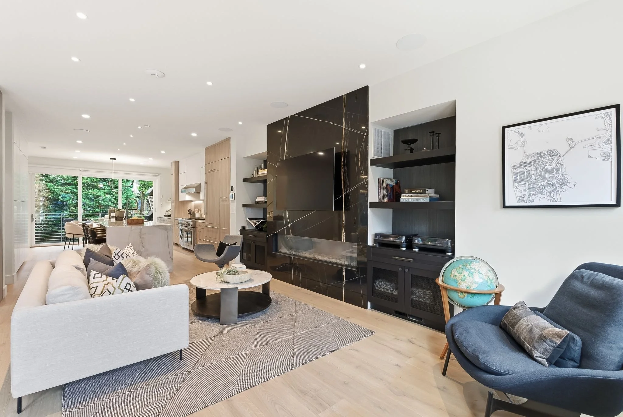 Modern living room with white sofa, patterned cushions, black marble fireplace, open kitchen area in background, and large window showing green trees outside.