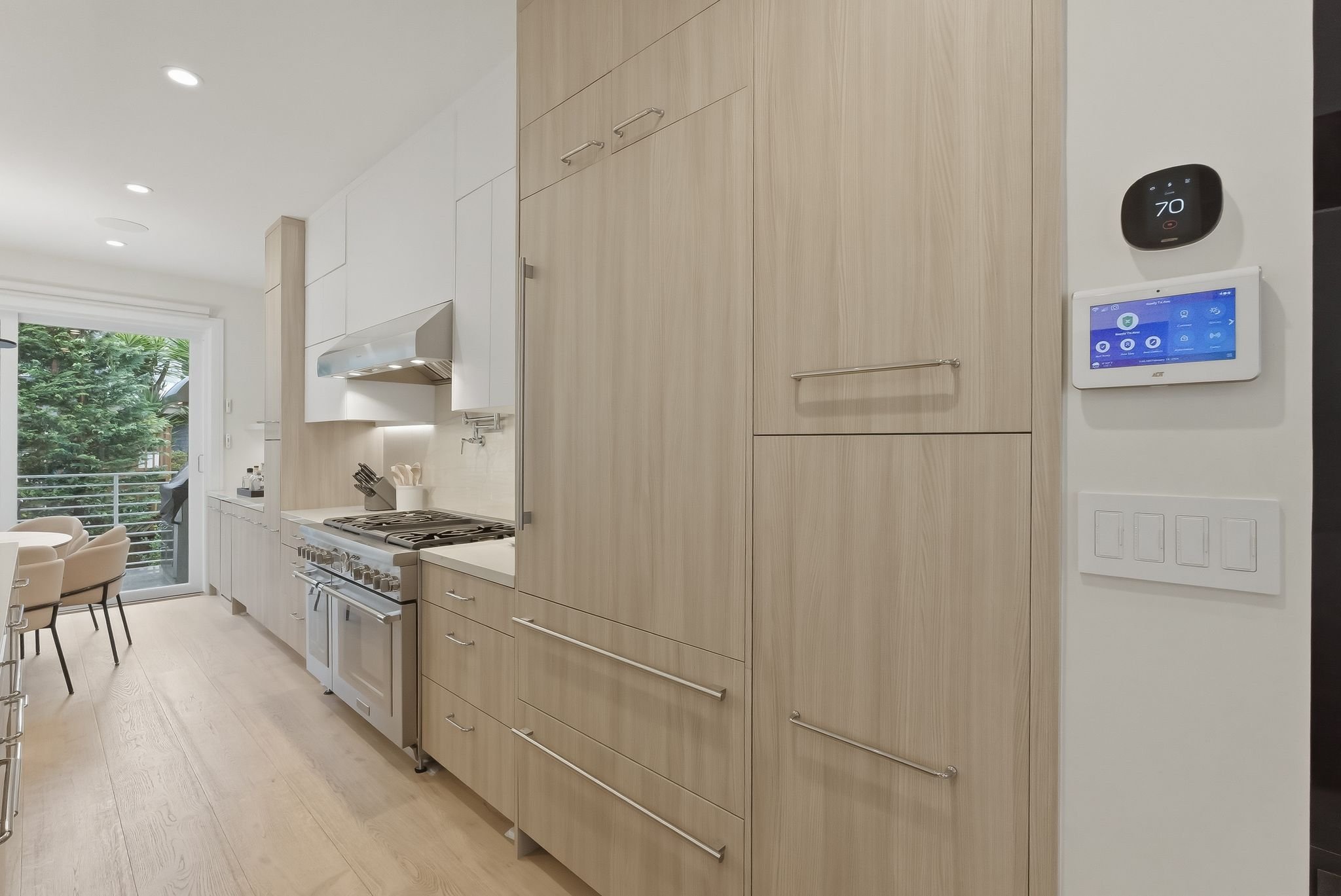 Modern kitchen with light wood cabinets, white countertops, a stainless steel stove, and a sliding glass door leading to a balcony with trees outside. Thermostat and smart home control panel are mounted on the wall.