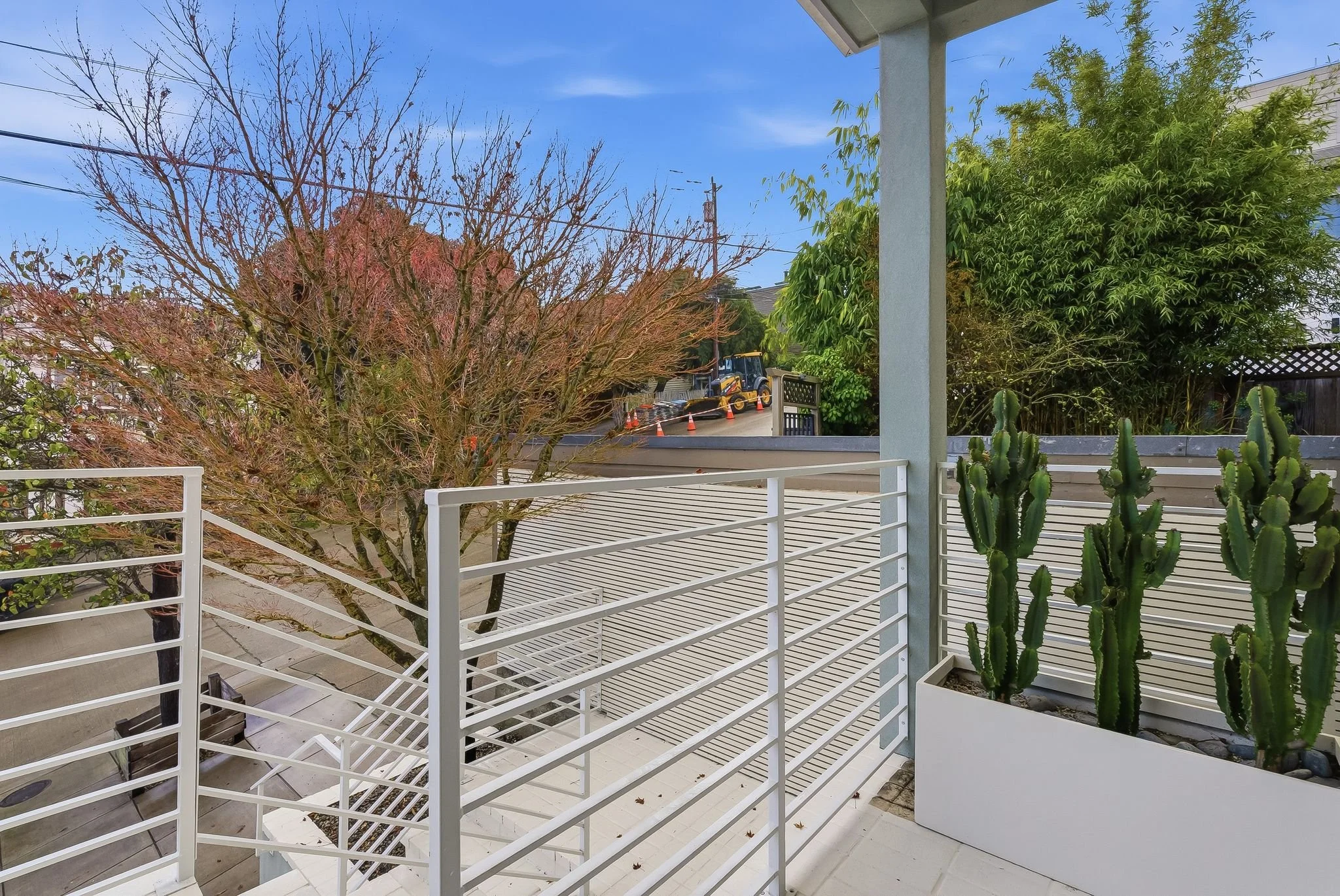 View from a balcony with white metal railing, potted cacti, and a large tree with sparse leaves; street and construction equipment with orange cones are visible beyond in a residential area with a blue sky.