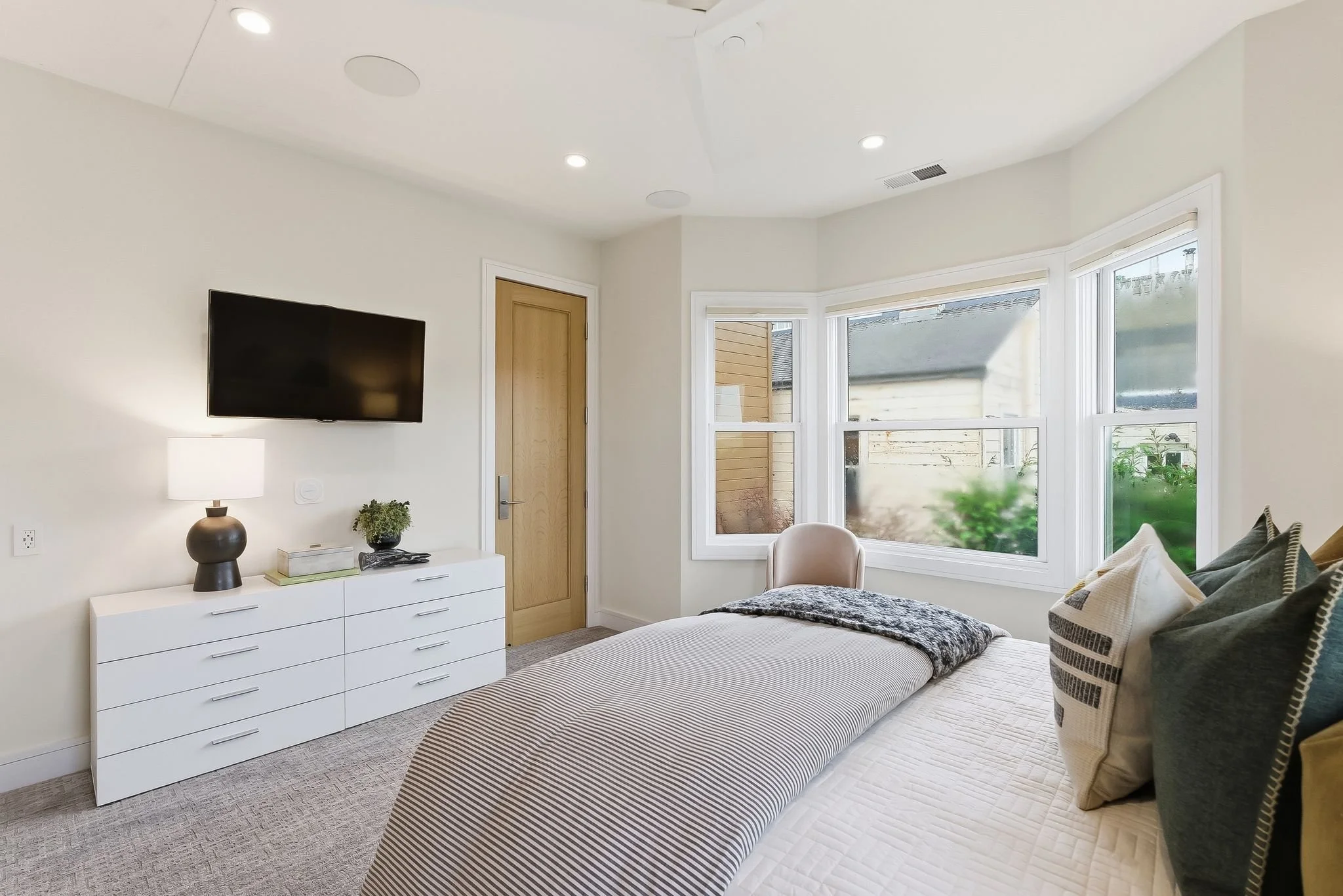 Bedroom with a bed, pillows, dresser with lamp and plant, flat-screen TV on wall, and a window showing outdoor view.