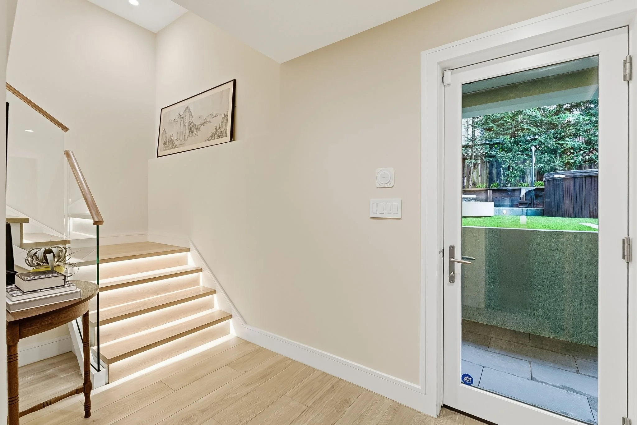 Interior view of a modern home entrance with a glass door leading outside to a backyard with a hot tub and greenery. Wooden stairs with LED lighting on each step and a small wooden table nearby.