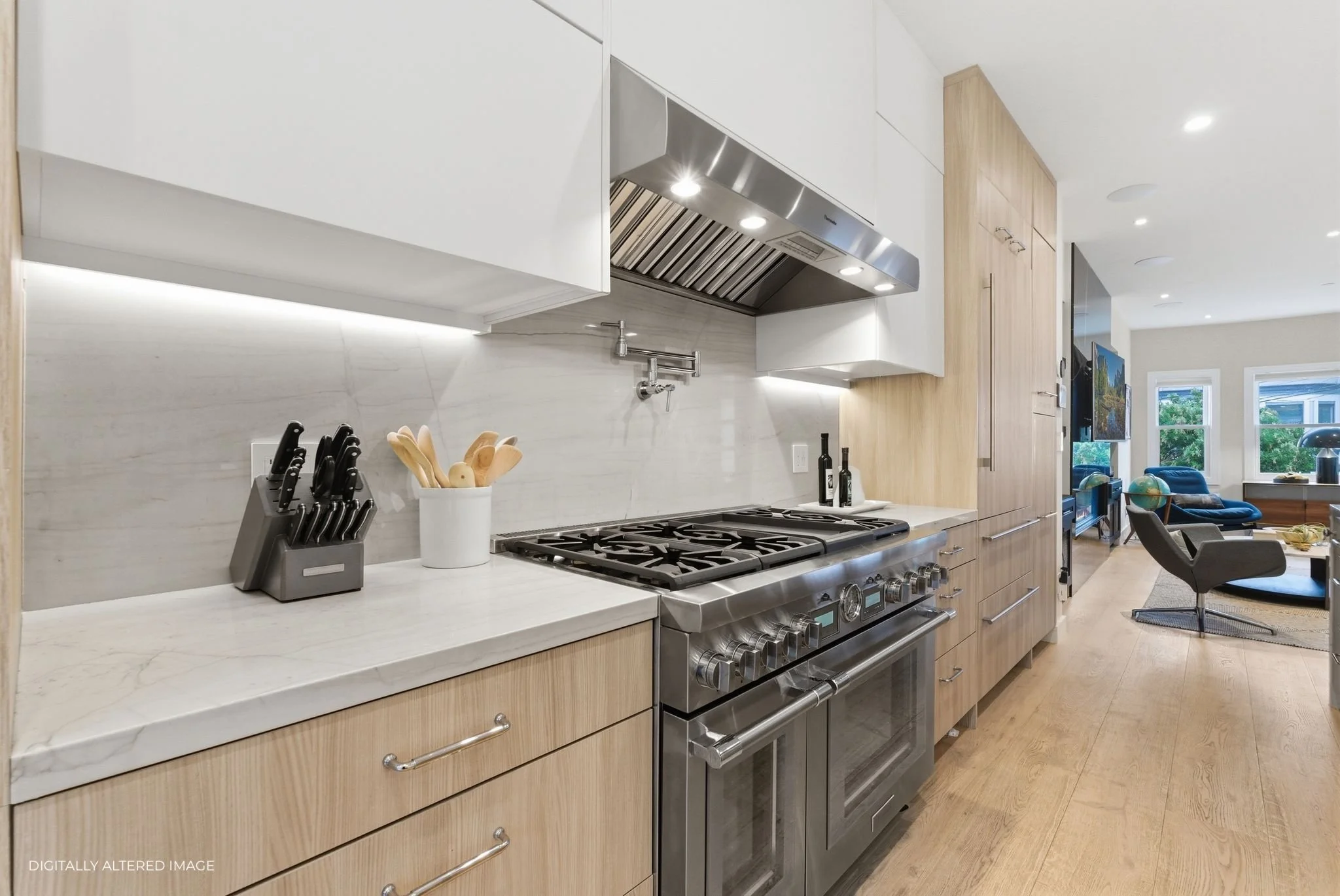 Modern kitchen with wooden cabinets, stainless steel stove, marble countertop, knife block, utensil holder, and view into living room with blue chairs and television.