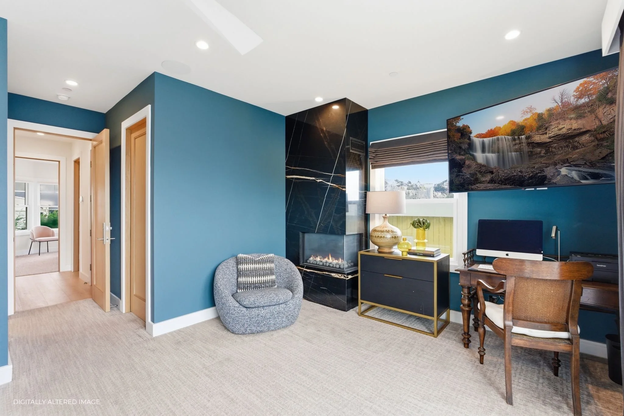 Living room with blue walls, a modern fireplace with black marble, a gray textured armchair, a table with a gold frame, lamp, and decorative items, a large wall-mounted TV displaying a waterfall, and a wooden desk with an iMac and office supplies.