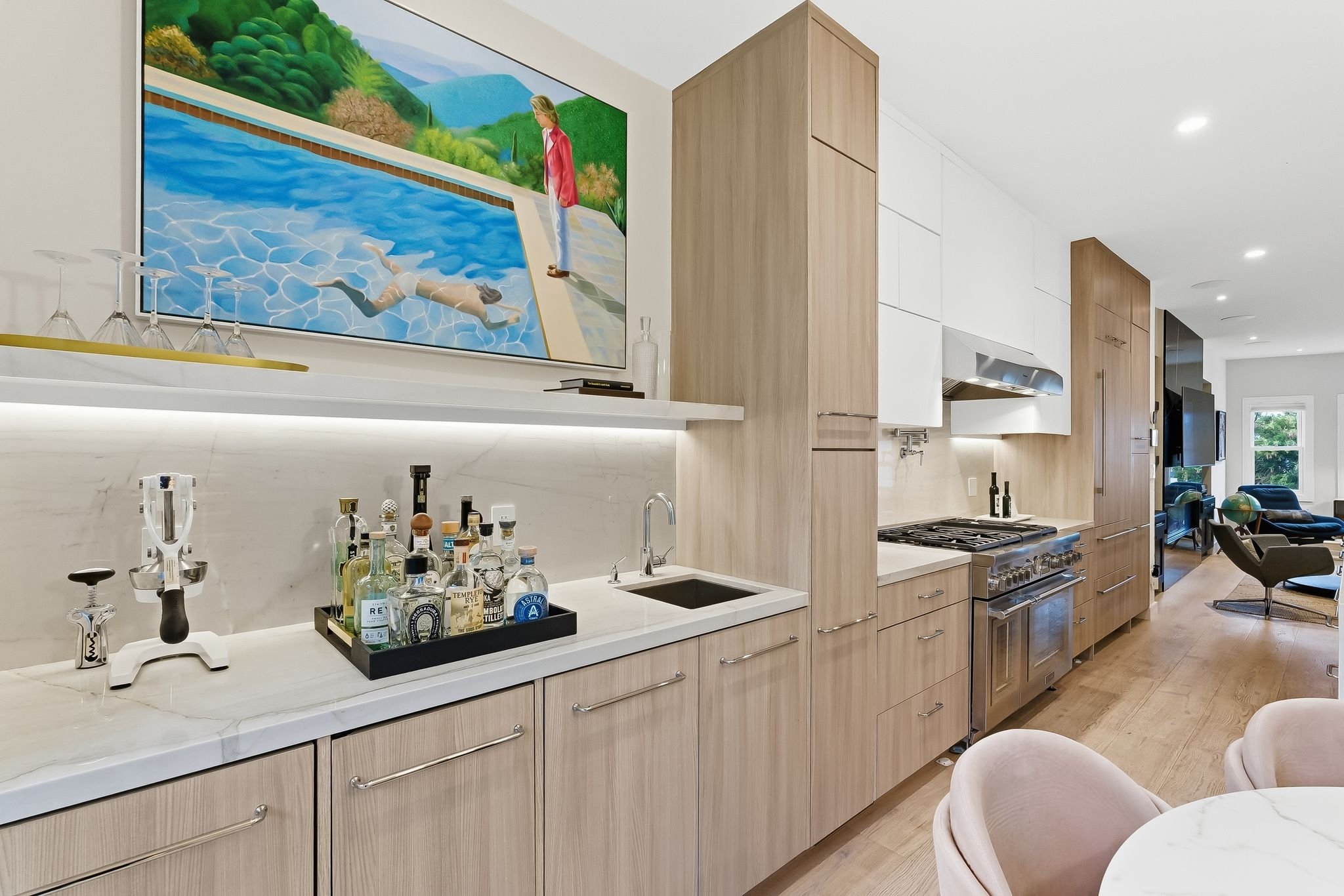Modern kitchen with light wood cabinets, white countertops, and a marble backsplash. A tray of liquor bottles is on the counter, and a sink is visible. There is a large abstract painting of a person swimming in a pool, with a woman observing from the