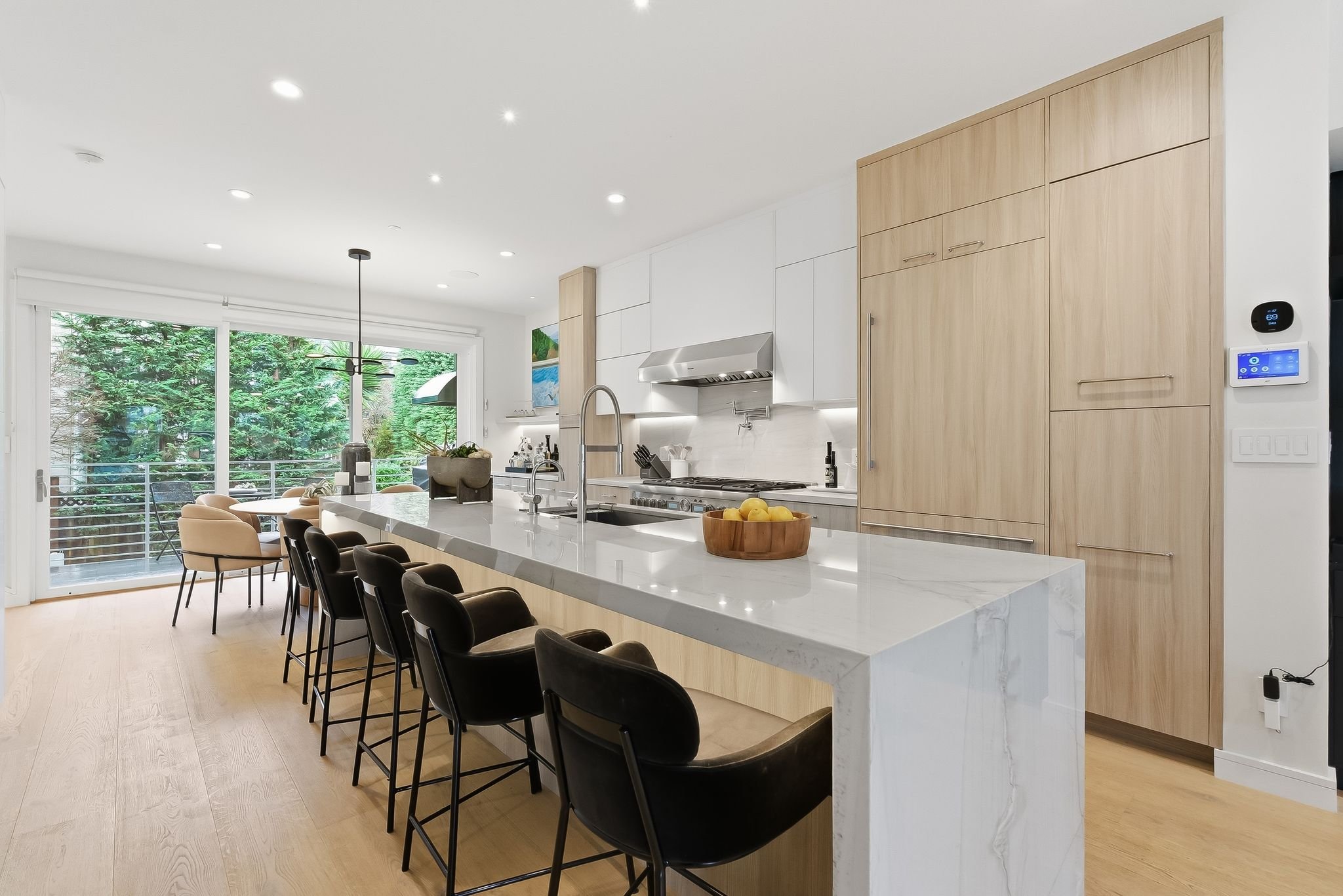 Modern kitchen with white walls, light wood cabinets, a large white marble island with black chairs, a sliding glass door leading to a deck, green trees outside, and minimalistic decor.