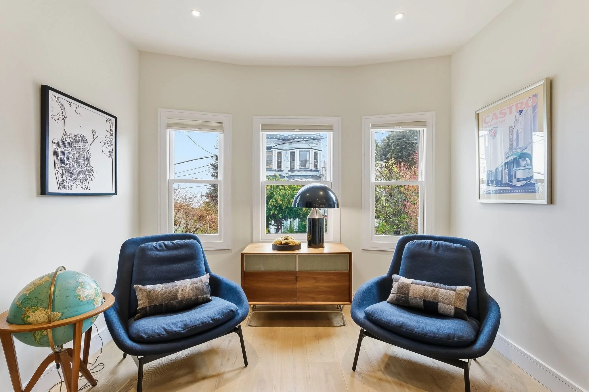 A cozy sitting room with two blue armchairs, a wooden sideboard with a black table lamp, a globe, and framed artwork on white walls, three windows showing a residential neighborhood.