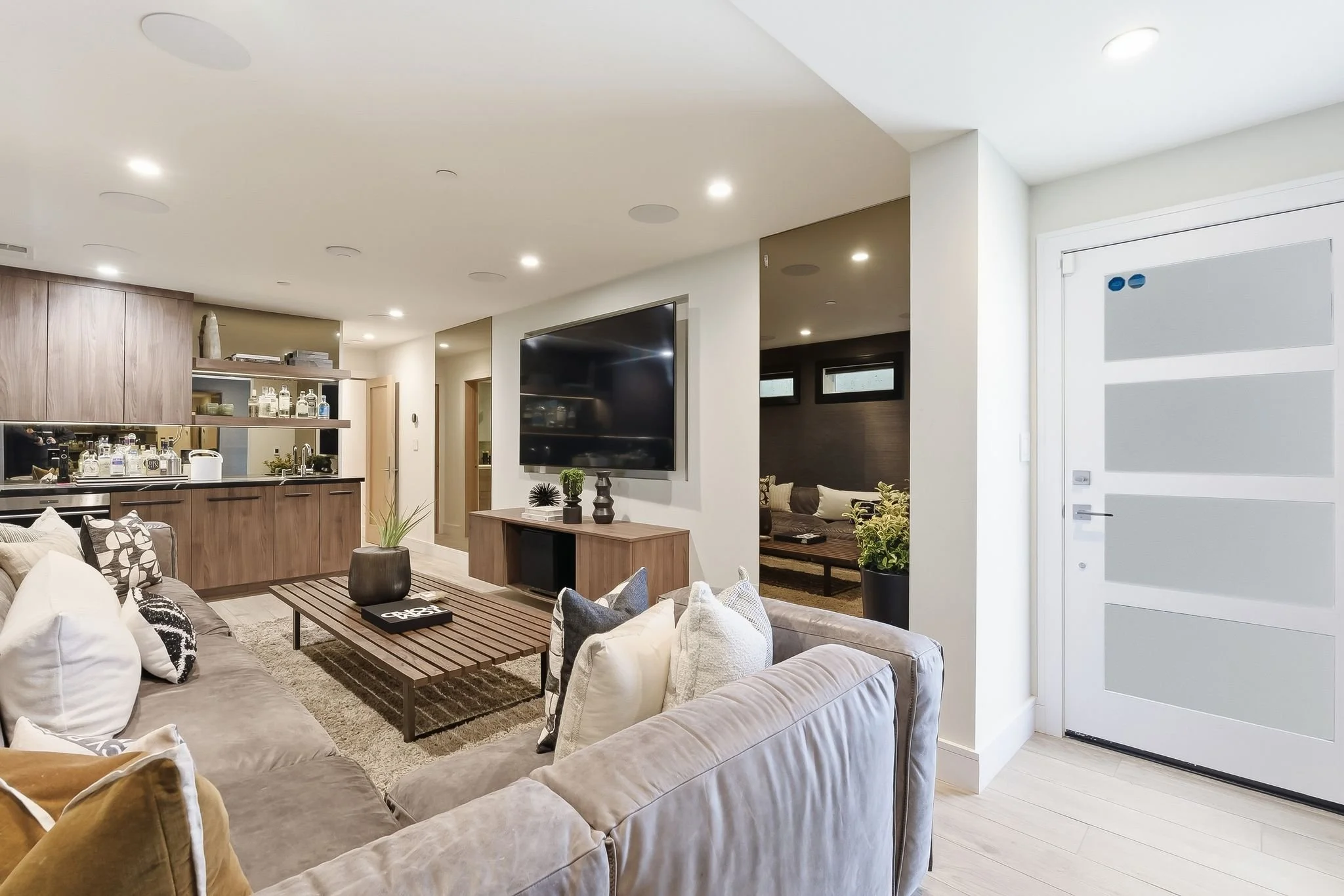 Modern living room with neutral-colored sofas, a wooden coffee table, and a wall-mounted flat-screen TV. The room has white walls, built-in ceiling lights, and a partial view of the adjacent kitchen with wooden cabinetry and open shelving. A white fr