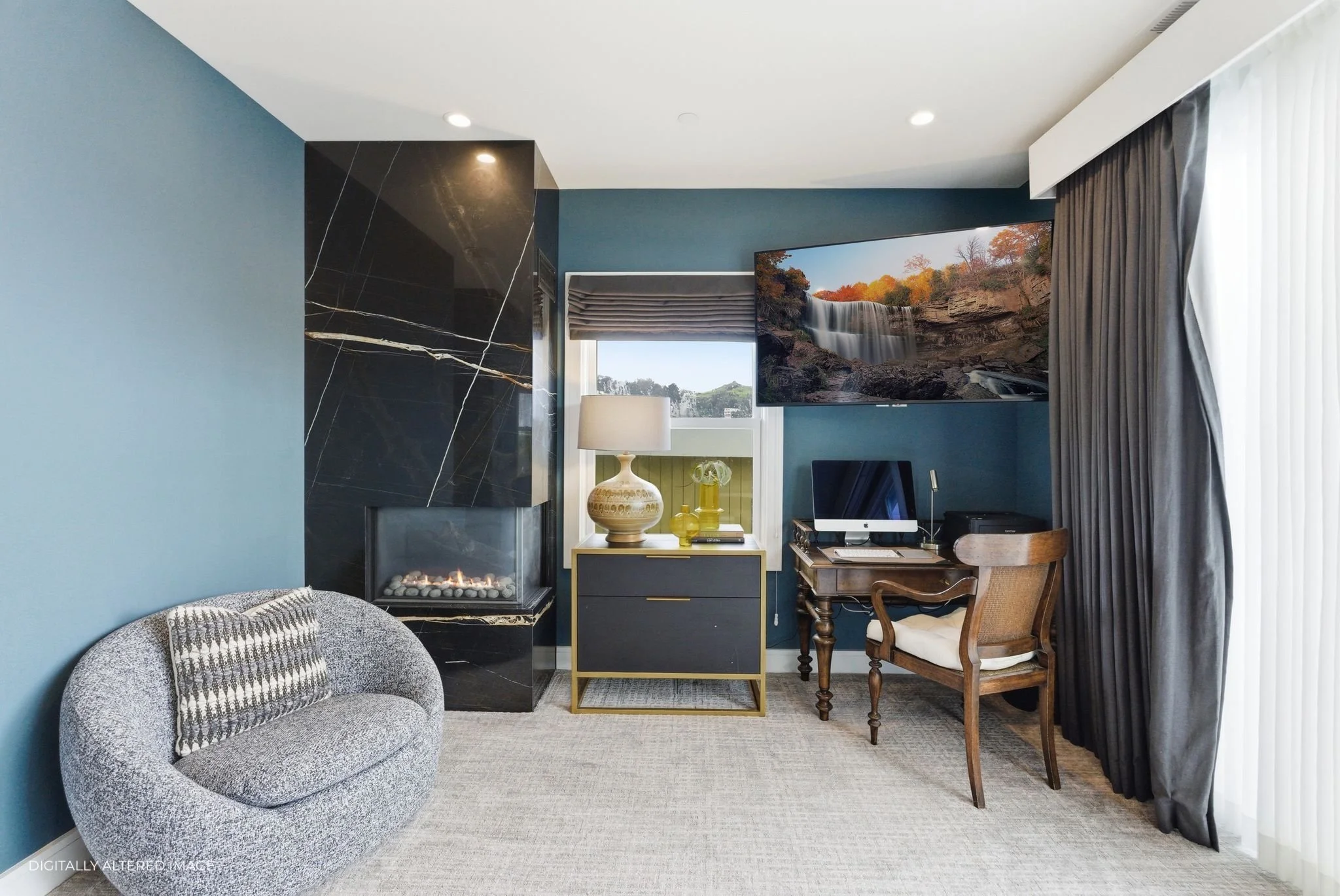 Living room corner with a small rounded gray sofa, fireplace with black marble and gold accents, a black side table with a white and beige vase, a window with a decorated shades, a desk with a computer and wooden chair, a large flat-screen TV. The ro