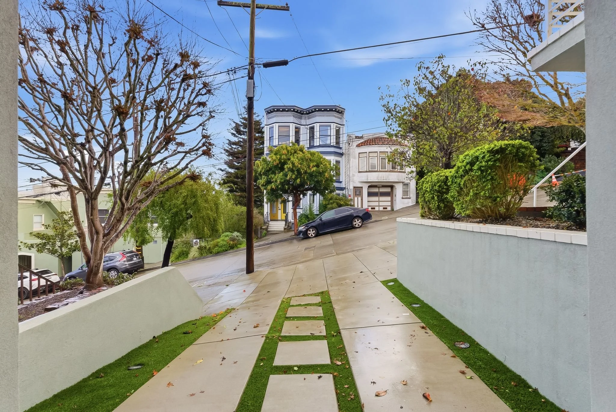 View of a residential neighborhood with a sloped intersection, trees, parked cars, and houses on a drizzly day.