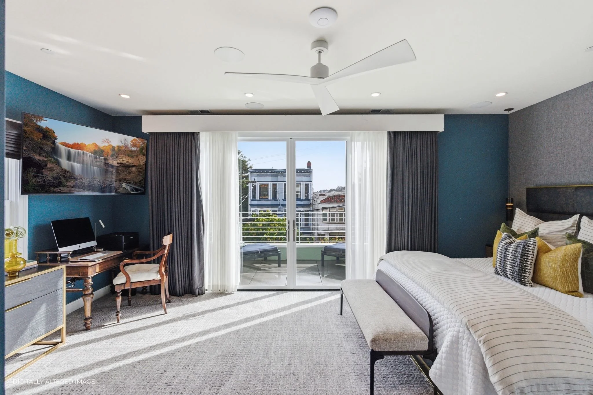 Brightly lit bedroom with a large bed, decorative pillows, and a balcony with outdoor seating accessible through sliding glass doors, a ceiling fan, and a wall-mounted flat-screen TV.