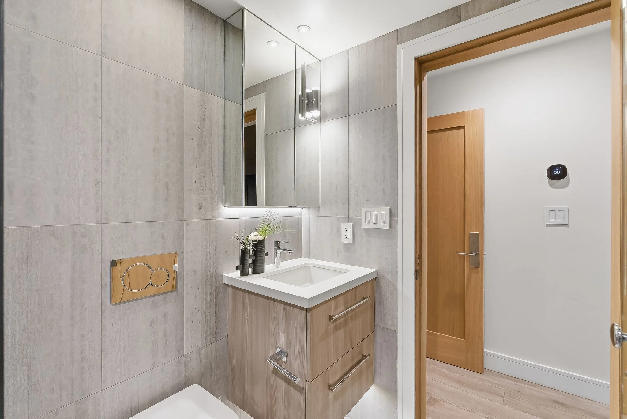 Modern bathroom with light wood vanity, white sink, mirror with backlight, wall-mounted black faucet, decorative black vases with flowers, wood and white door, digital thermostat on white wall, and light gray tiled walls.