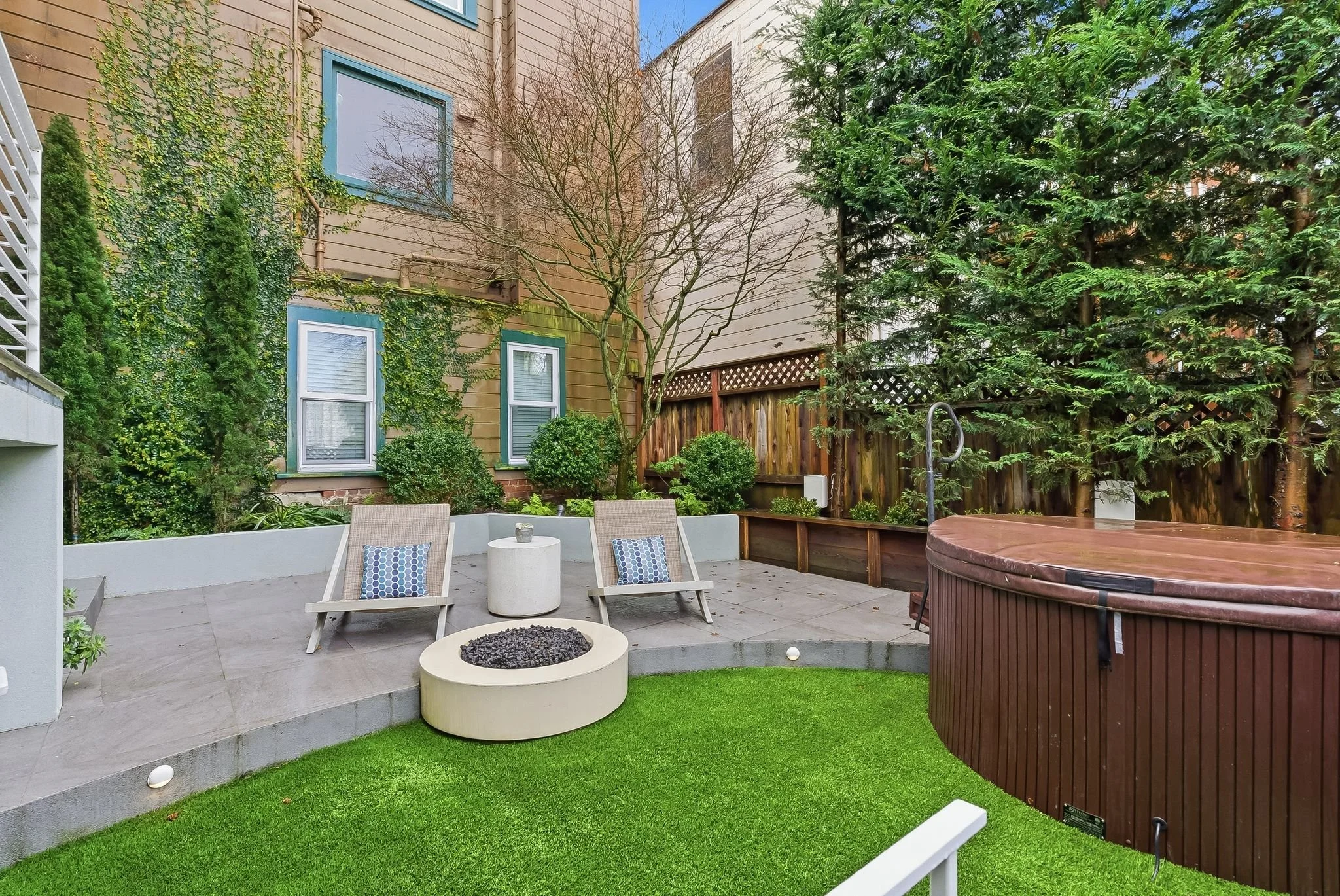A backyard patio with two lounge chairs and a small table on a tiled surface, next to a fire pit, with a grassy area in the foreground. There is a hot tub to the right, and a raised garden bed with bushes and trees along a wooden fence in the backgro