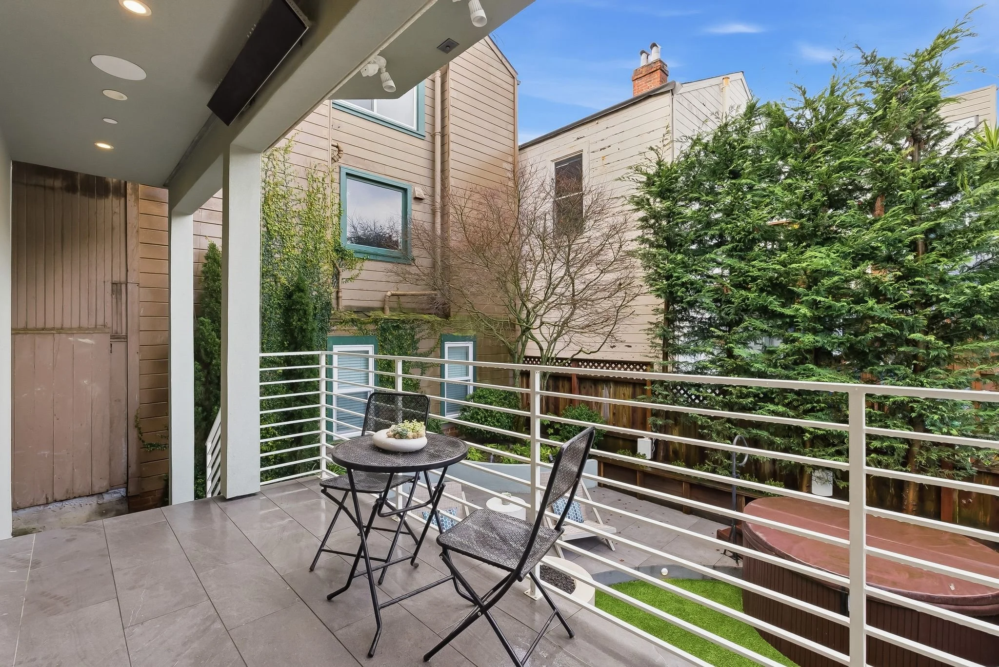 Balcony with two black chairs and a small round table with a white decorative bowl, overlooking a backyard with a hot tub, lounge chairs, trees, and neighboring houses under a blue sky.