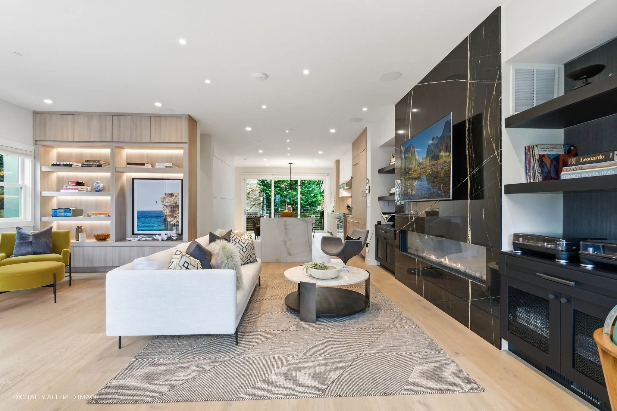 Modern living room with white sofa, yellow accent chair, black marble fireplace with TV, built-in shelving, and a view into the kitchen and dining area with large windows showing green trees outside.