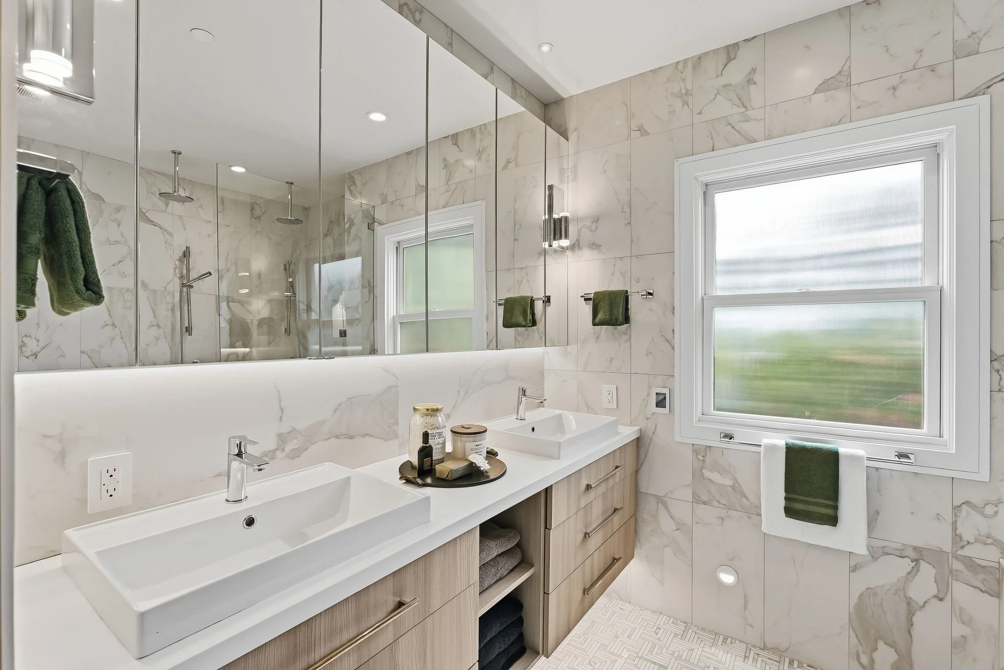 Modern bathroom with double vanity, marble walls, large window, and walk-in shower with glass doors and rain showerheads.