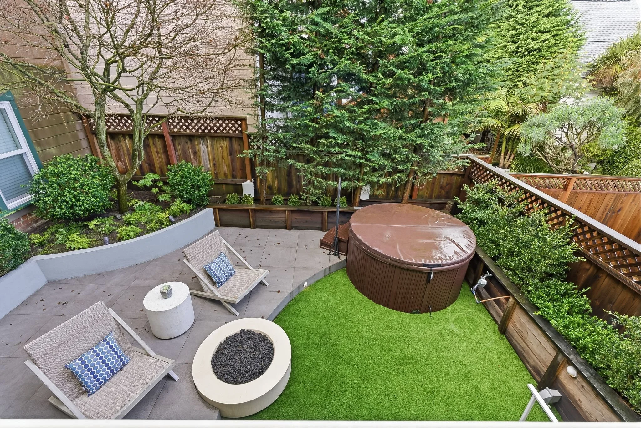 A backyard patio with two beige lounge chairs with blue and white pillows, a round white fire pit with black stones, a hot tub covered with a brown lid, and greenery including trees, shrubs, and a well-manicured lawn separated by a wooden fence.