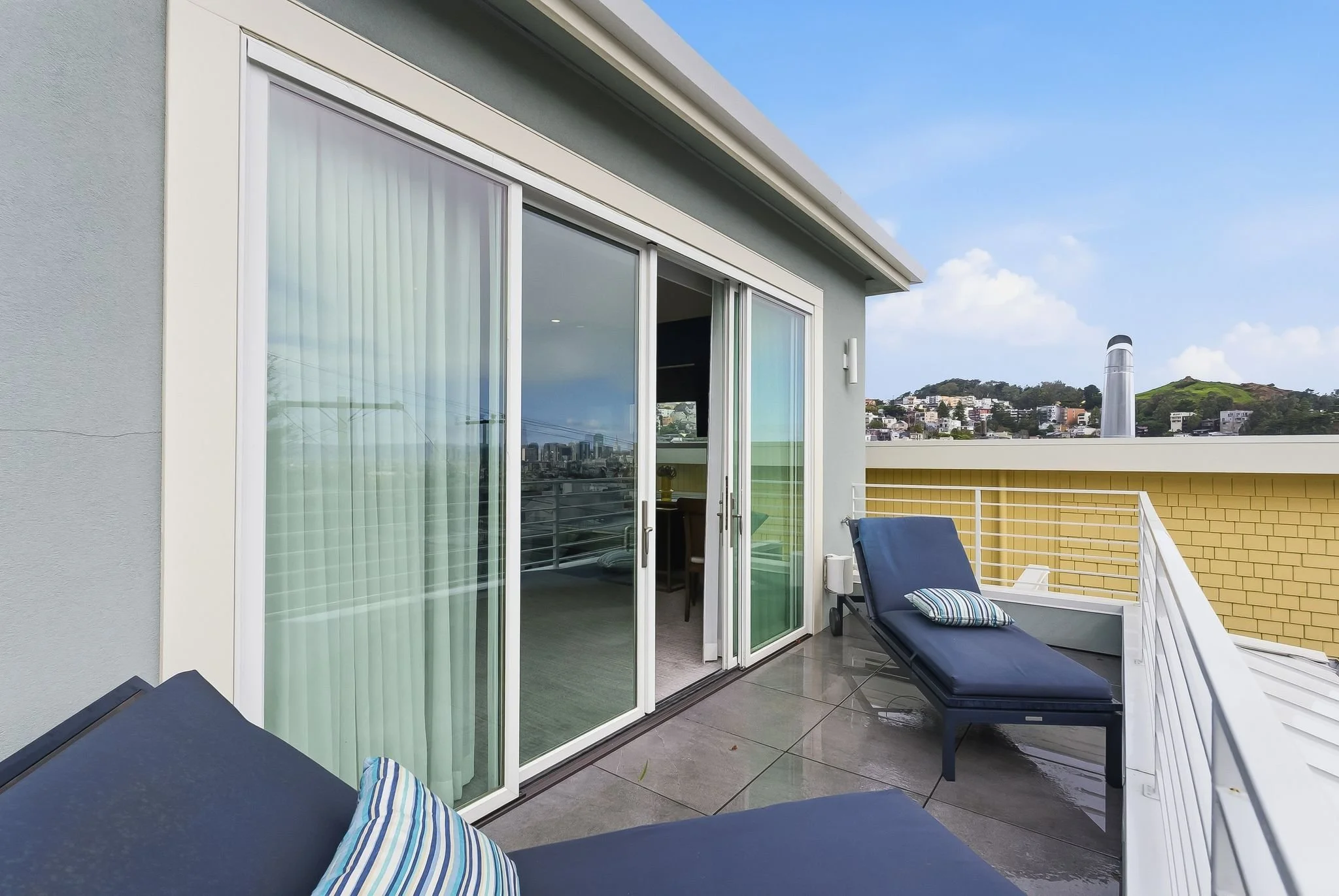 A modern rooftop balcony with black lounge chairs, striped pillows, sliding glass doors, a view of the city skyline, hills, and a partly cloudy sky.