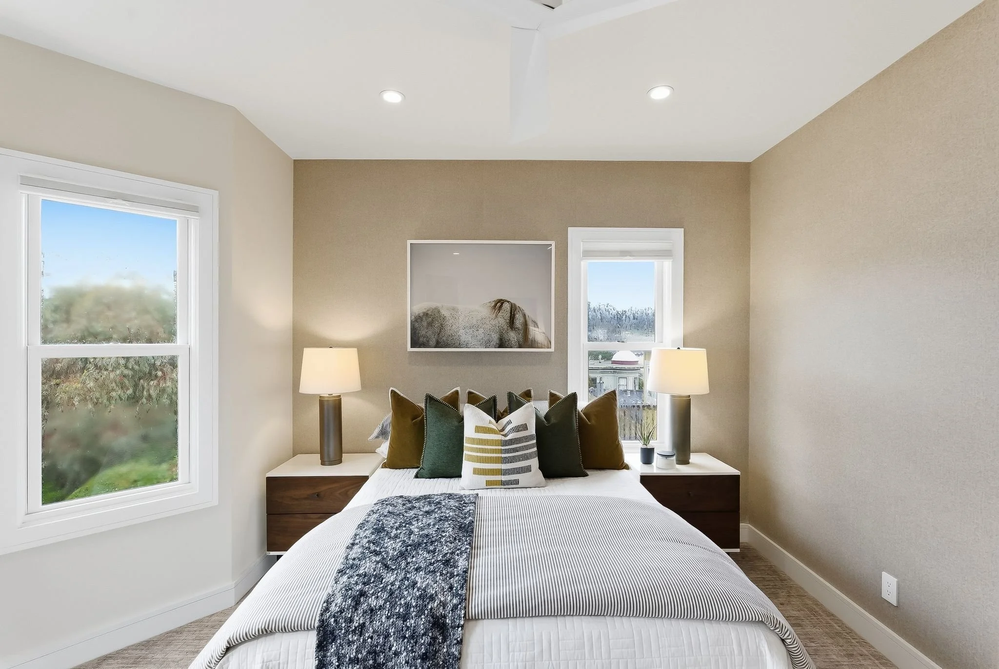 A cozy bedroom with beige walls, a large window on each side of the bed, two lamps on bedside tables, various pillows on the bed, and a black-and-white artwork above the headboard.