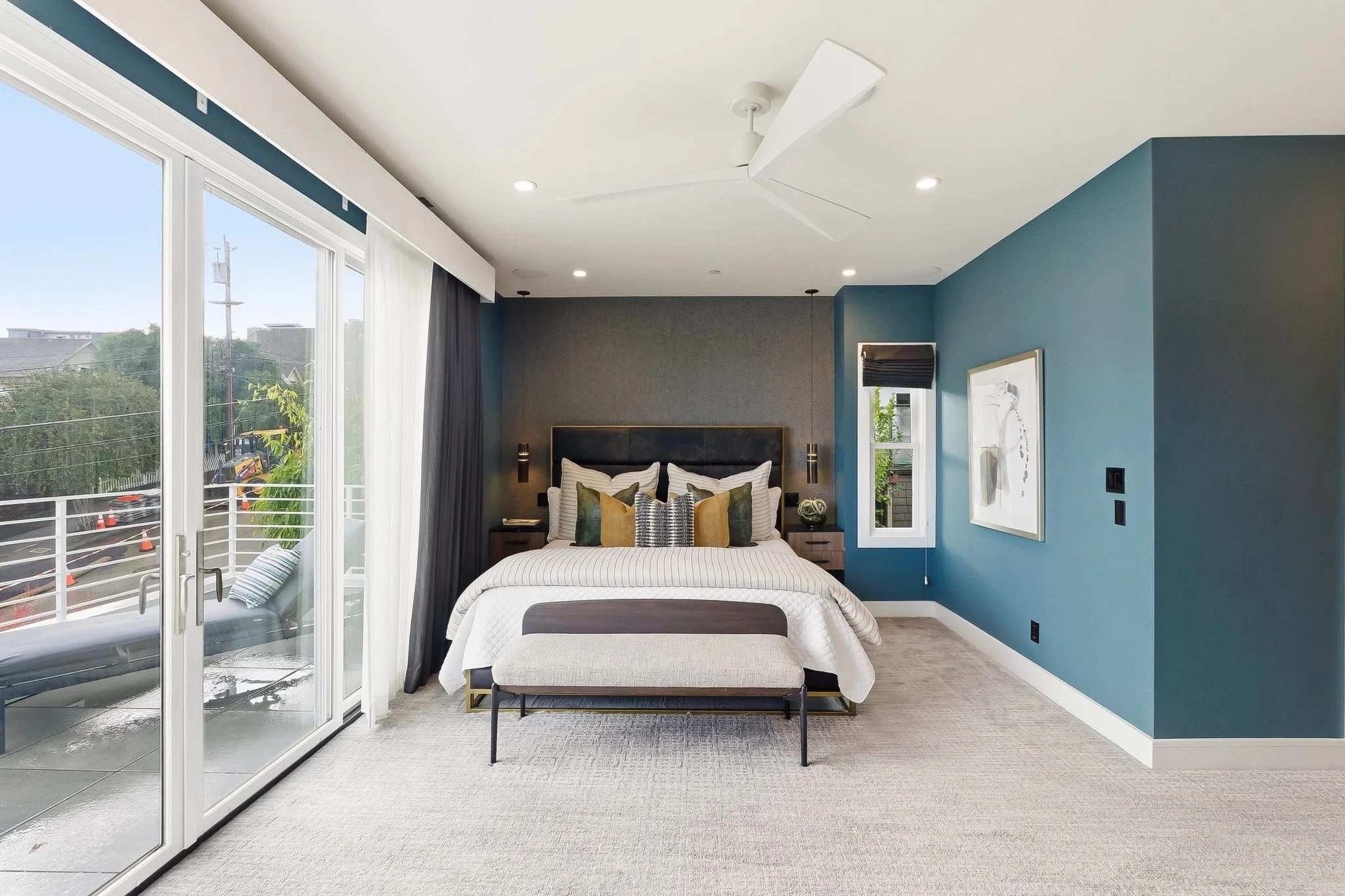 Modern bedroom with large sliding glass door, king-sized bed with multiple pillows, a bench at the foot of the bed, wall art, and a ceiling fan.