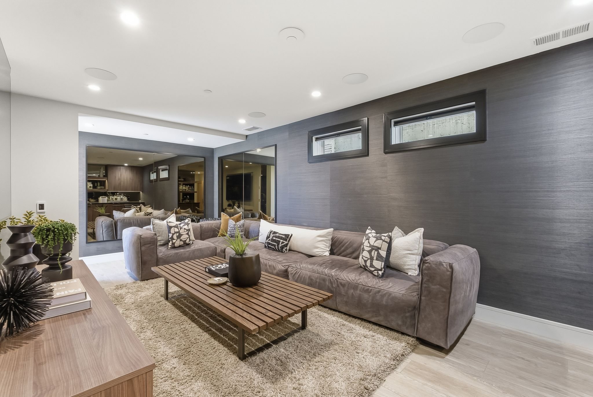 Modern living room with a large gray sectional sofa, decorative pillows, wooden coffee table, plants, and a dark textured accent wall with windows.