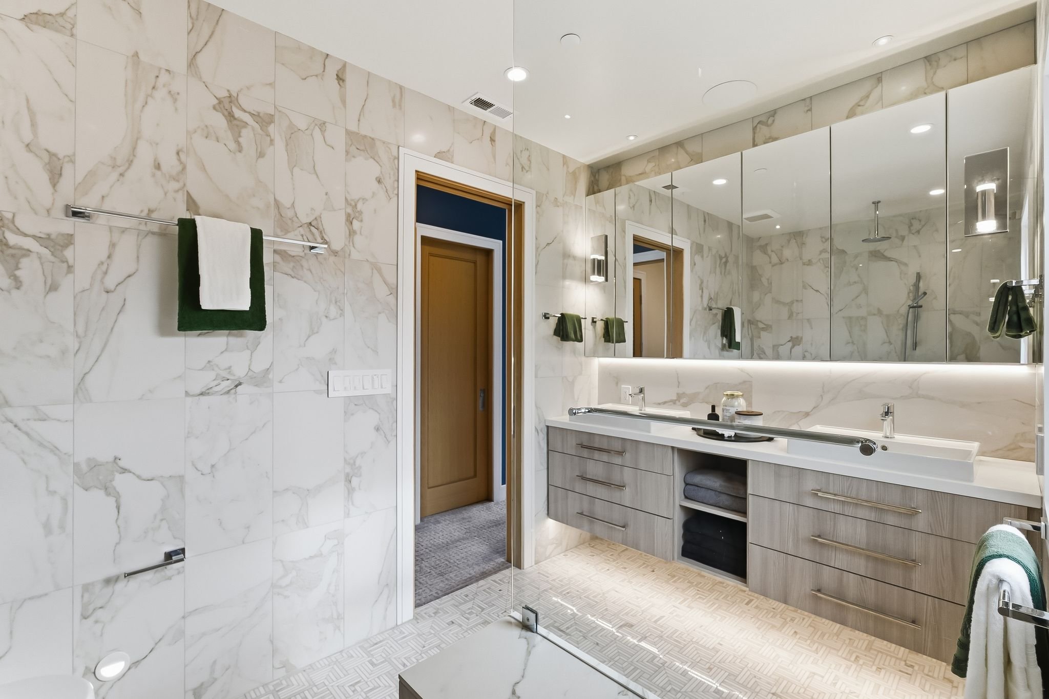 Modern bathroom with marble walls, a large mirror, double sink vanity, towel racks with towels, and a glass shower enclosure.