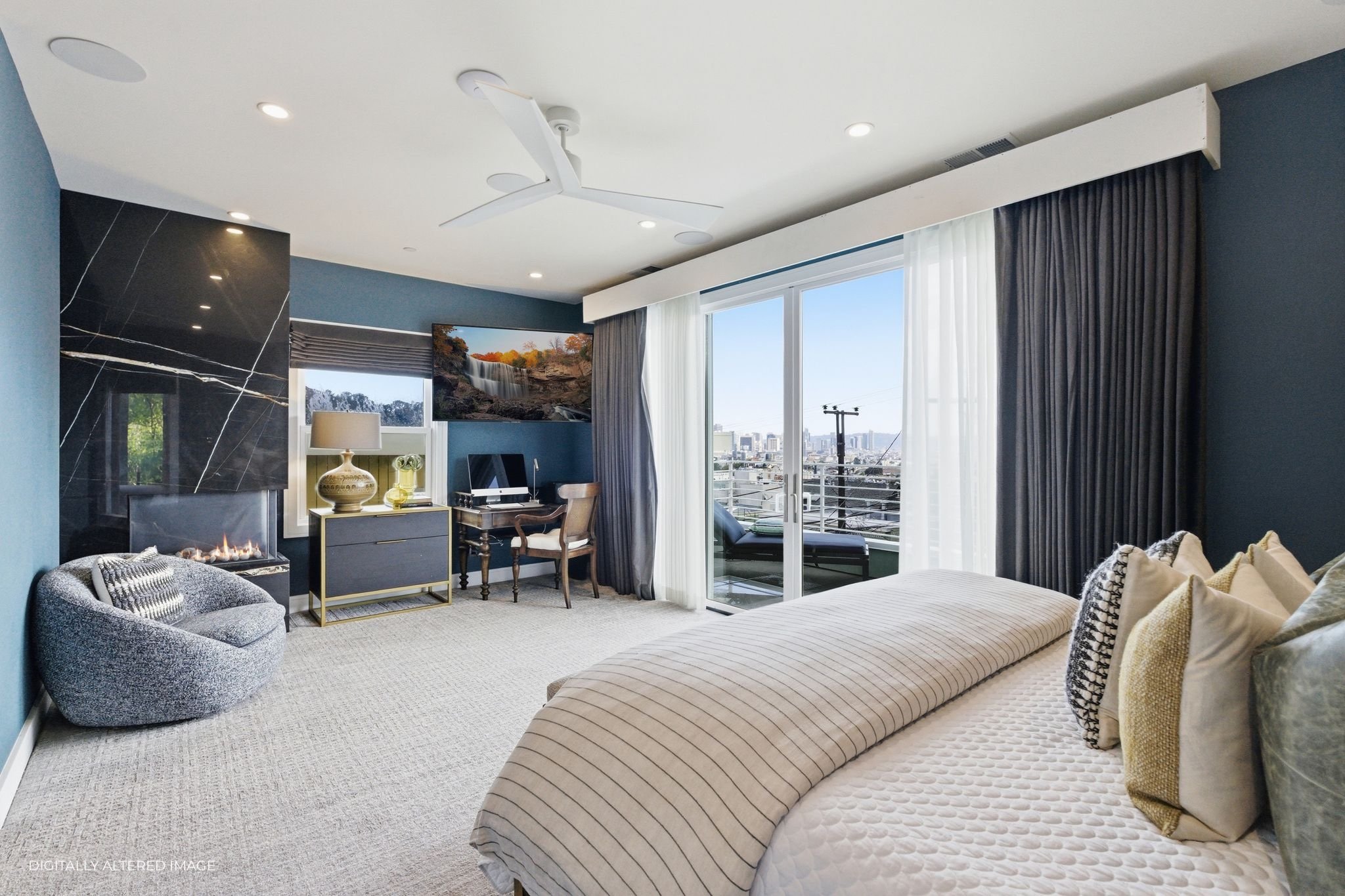 Modern bedroom with a bed, a sitting chair by a fireplace, a desk with a chair, a large window with curtains, a balcony, and city view