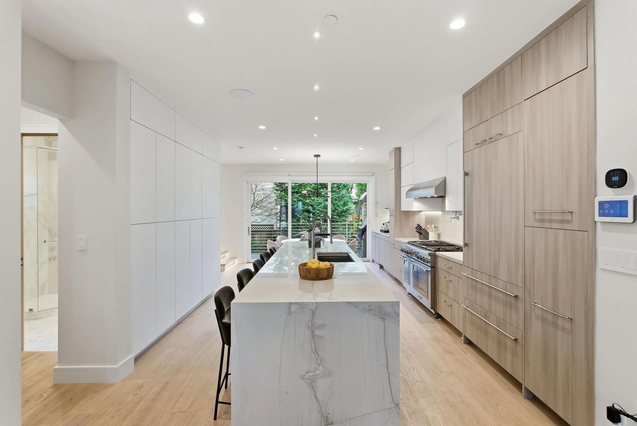 Modern kitchen with white marble island, black chairs, light wood cabinetry, stainless steel appliances, and a sliding glass door leading to an outdoor area.