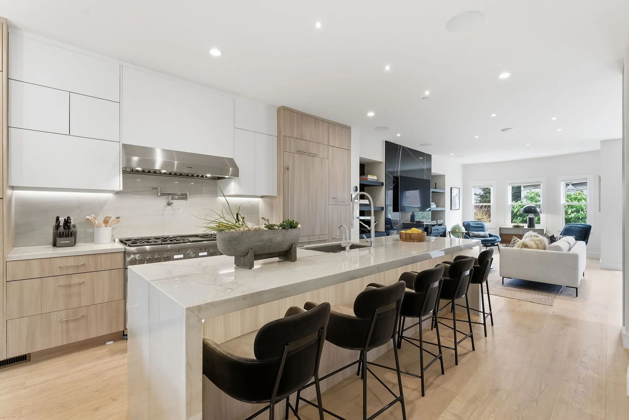 Open-concept kitchen and living room with minimalist decor, white cabinets, light wood accents, black chairs, and large windows with a view of greenery.