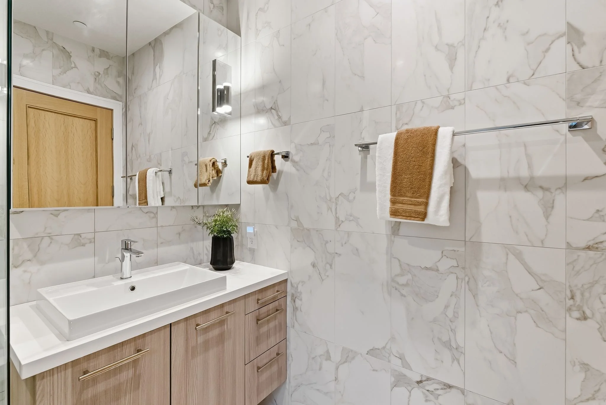 Modern bathroom with a marble wall, wooden vanity with a rectangular white sink, towel bars with brown and white towels, a mirror, and a small black vase with green plant.