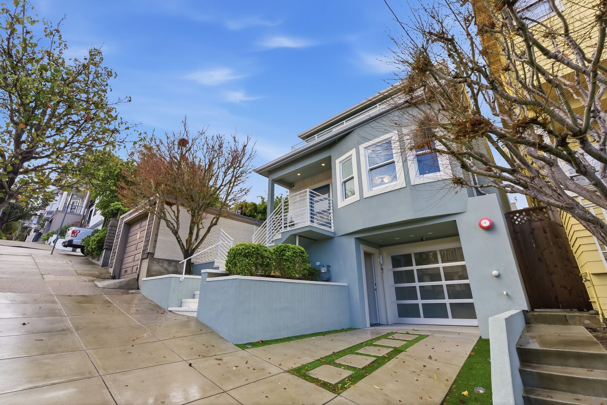 Modern multi-story house with blue exterior, balcony, and garage, surrounded by bare and leafy trees on a sloped street.