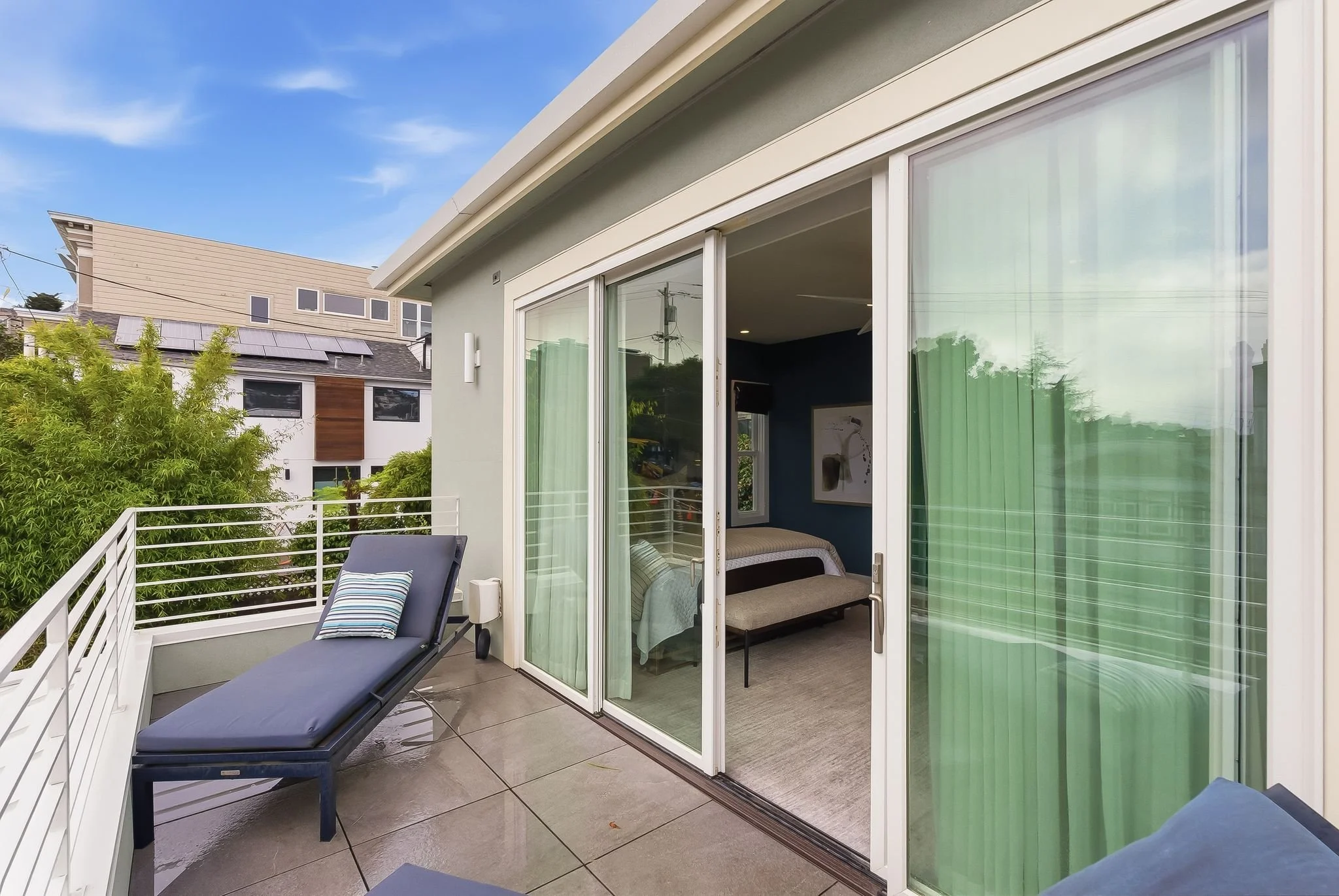 View of a balcony with two lounge chairs, one with a striped pillow, and a sliding glass door leading into a bedroom with dark blue walls and a bed.