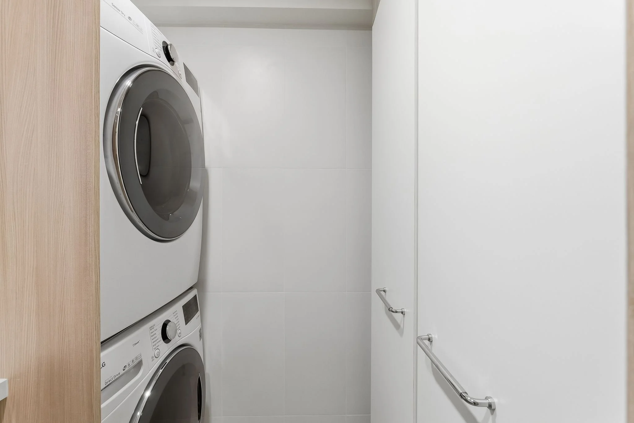 A small laundry room with a front-loading washing machine and dryer stacked against the wall, with white tiled walls and two metal grab bars on the right wall.