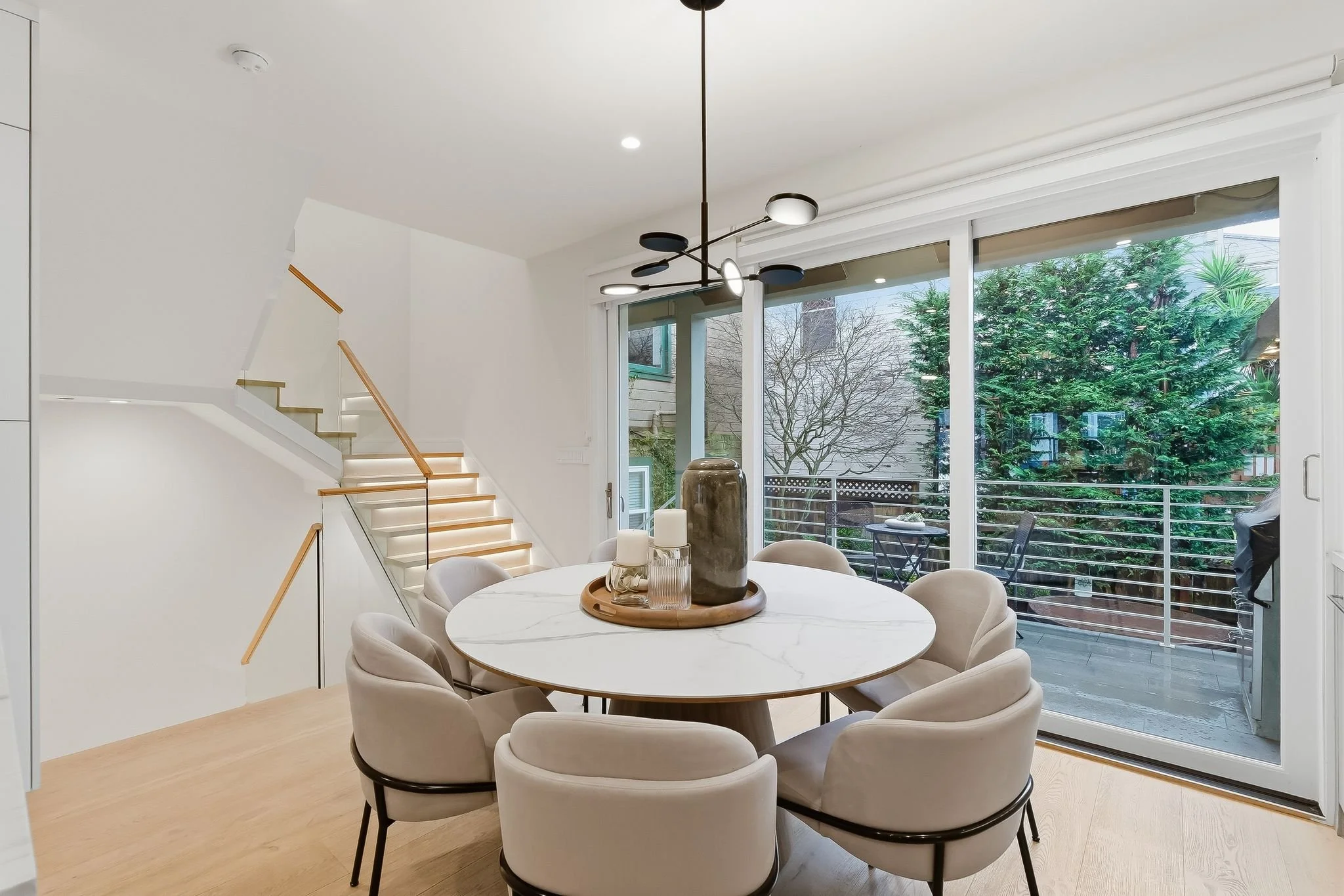 Modern dining room with round white marble table surrounded by eight beige upholstered chairs, large sliding glass door leading to an outdoor balcony with trees and railing, staircase with wooden handrail and glass panels to the left, minimalist blac