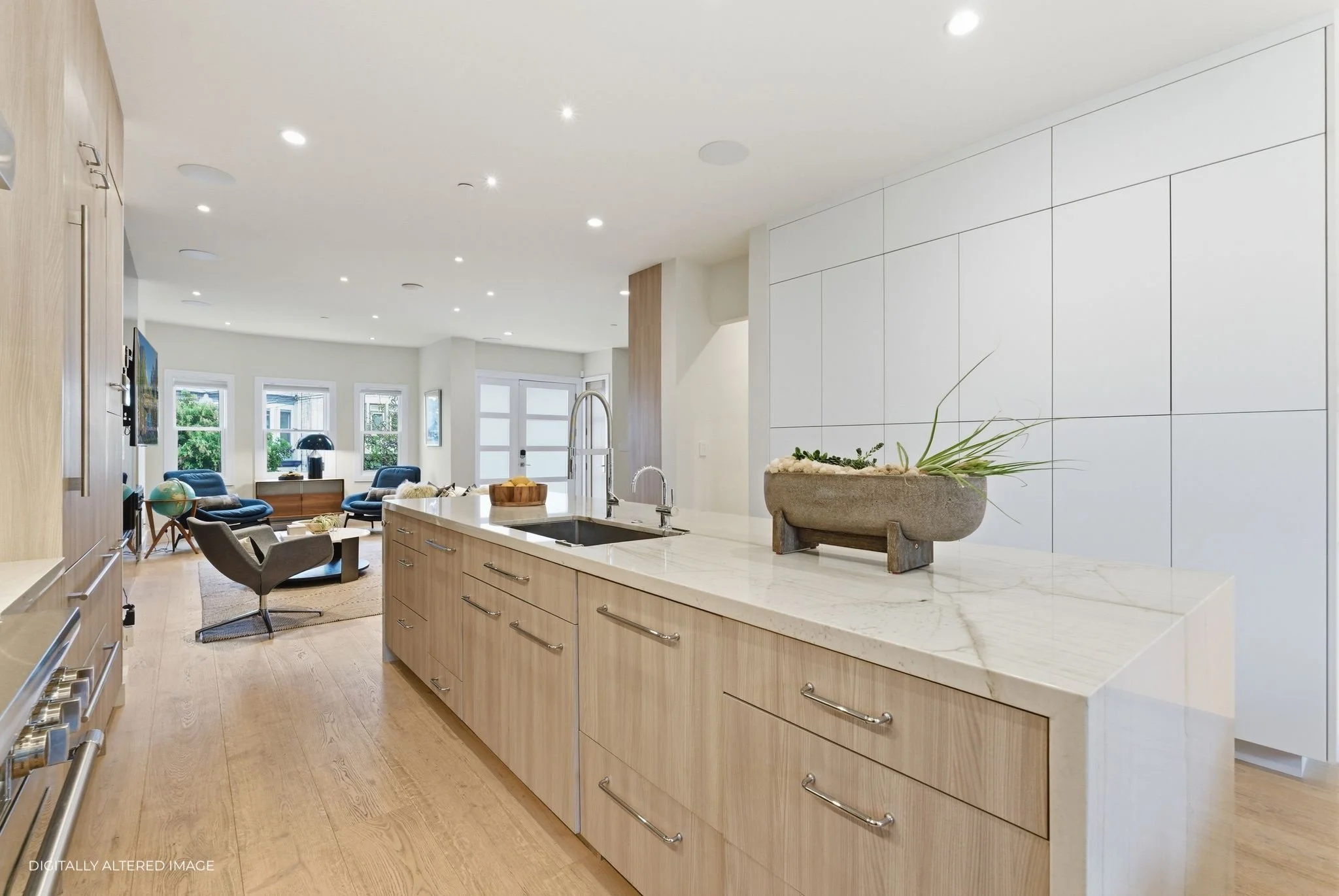 Open-concept kitchen with a marble countertop island, light wood cabinets, and a view into a bright living room with large windows and modern furniture.