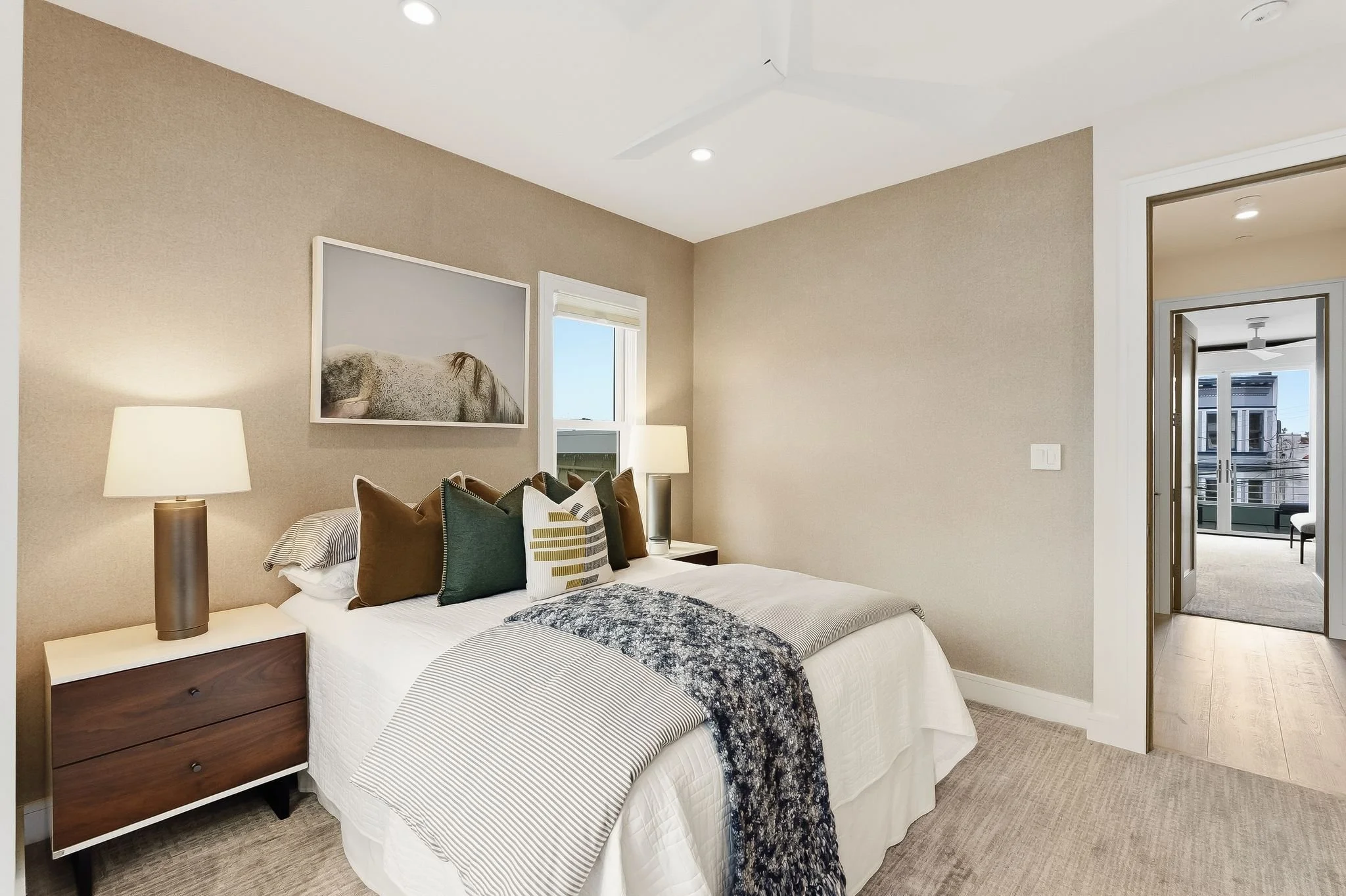 A bedroom with a bed featuring multiple pillows, a nightstand with a lamp, a framed picture of a horse above the bed, a window, and an adjoining room with sliding glass doors opening to a balcony.