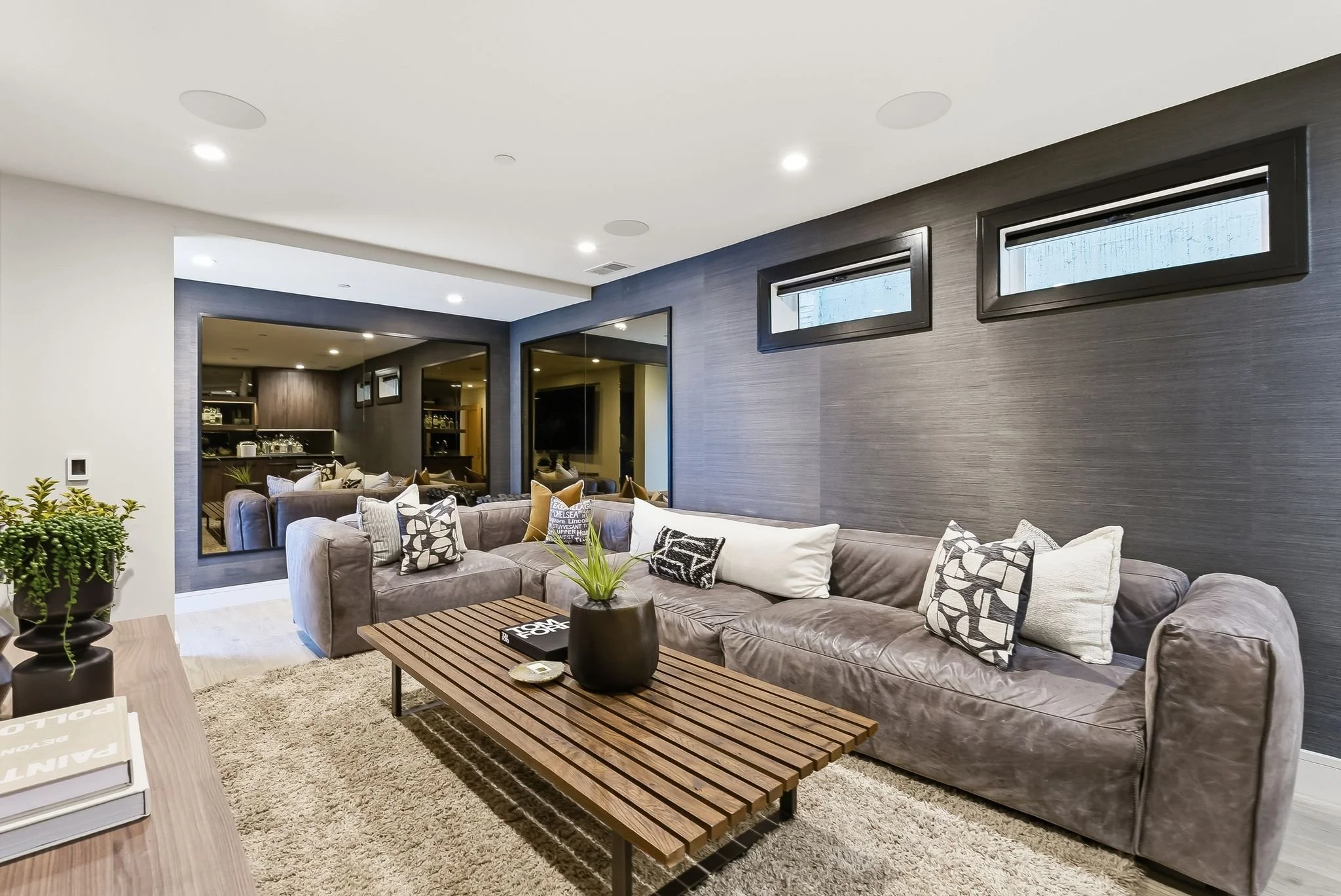 Modern living room with gray sectional sofa, wooden coffee table, patterned pillows, and dark textured accent wall, with small rectangular windows above.
