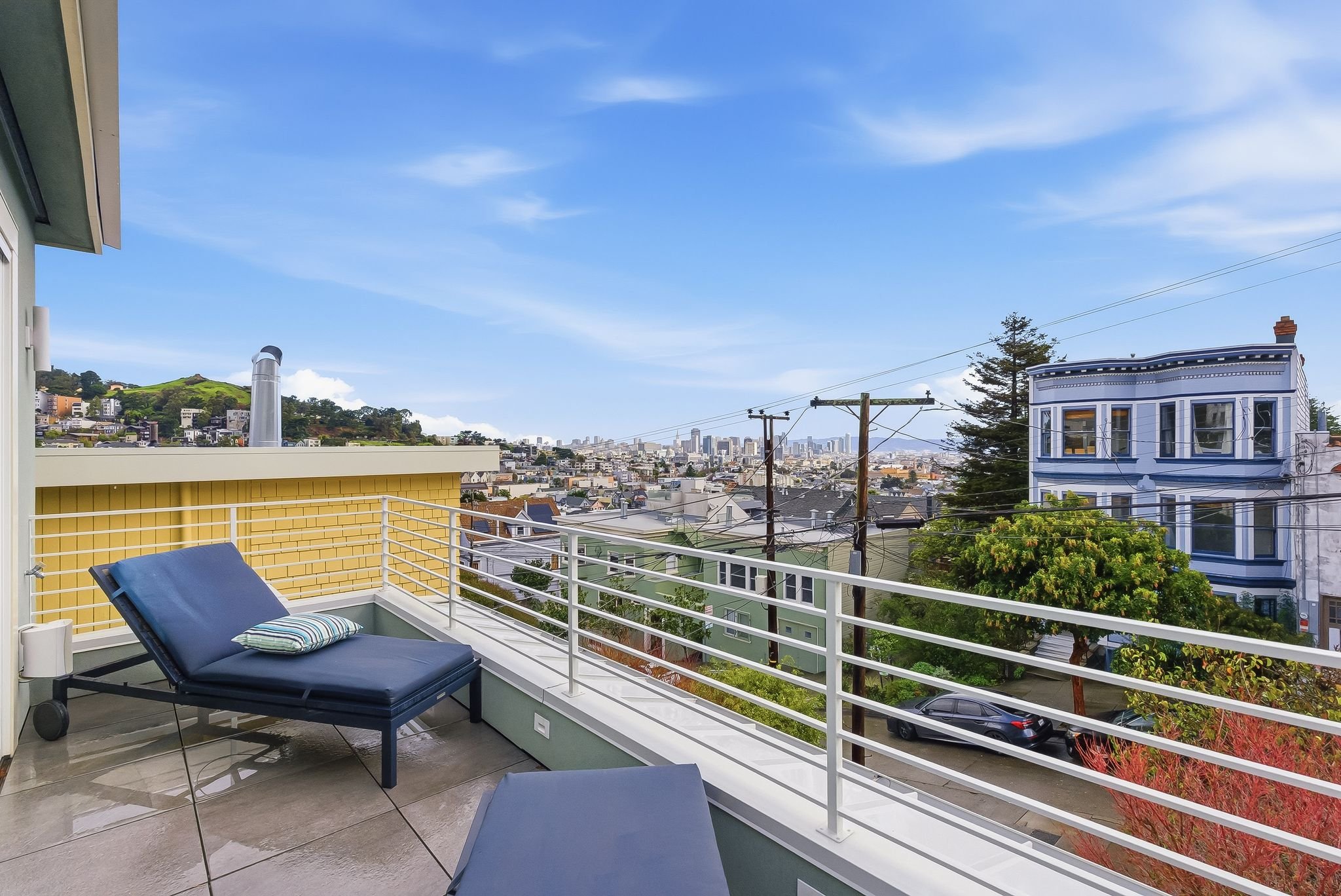 Balcony with a blue lounge chair and pillow, overlooking a cityscape with colorful buildings, green trees, and a blue sky.