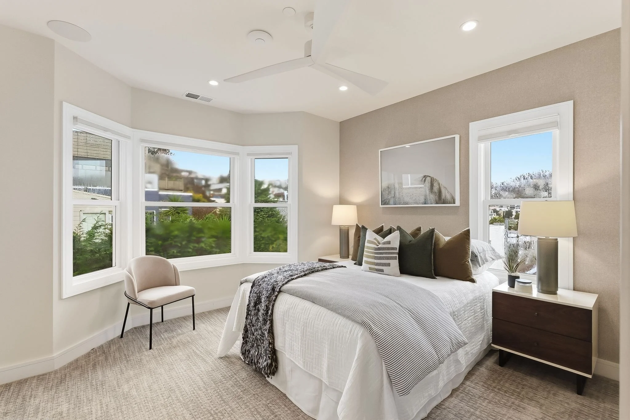 Bright bedroom with a bed, nightstands with lamps, a chair, and large bay windows showing greenery outside.