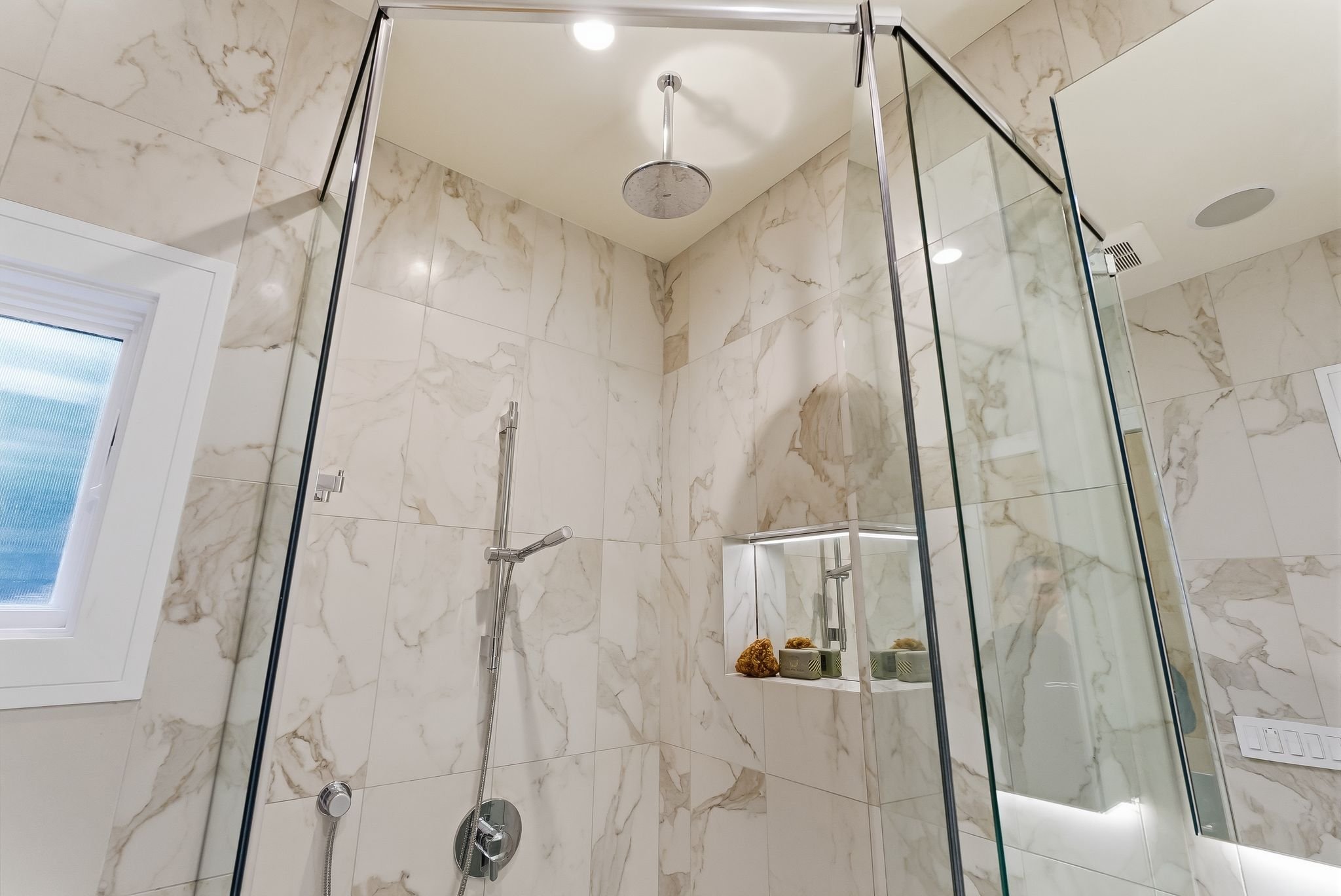 A modern shower with glass enclosure, marble wall tiles, a ceiling rain shower head, a handheld shower, a small built-in shelf with decorative stones, and a window with frosted glass.
