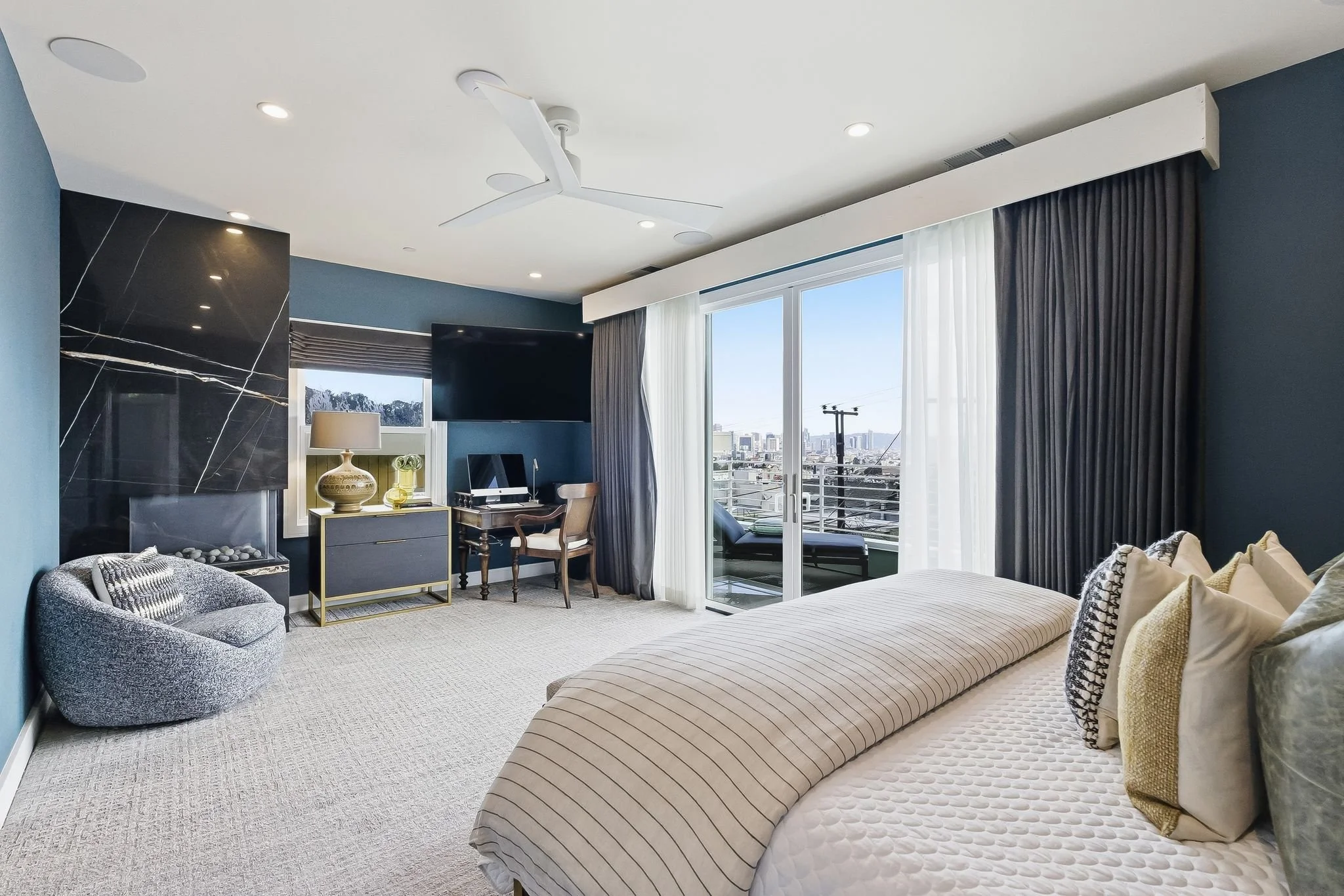 Modern bedroom with a large bed, decorative pillows, and a rolled-up blanket. A sliding glass door leads to a balcony with city views. There is a flat-screen TV, a desk with a chair, a black fireplace, a gray armchair, and a lamp with a decorative va