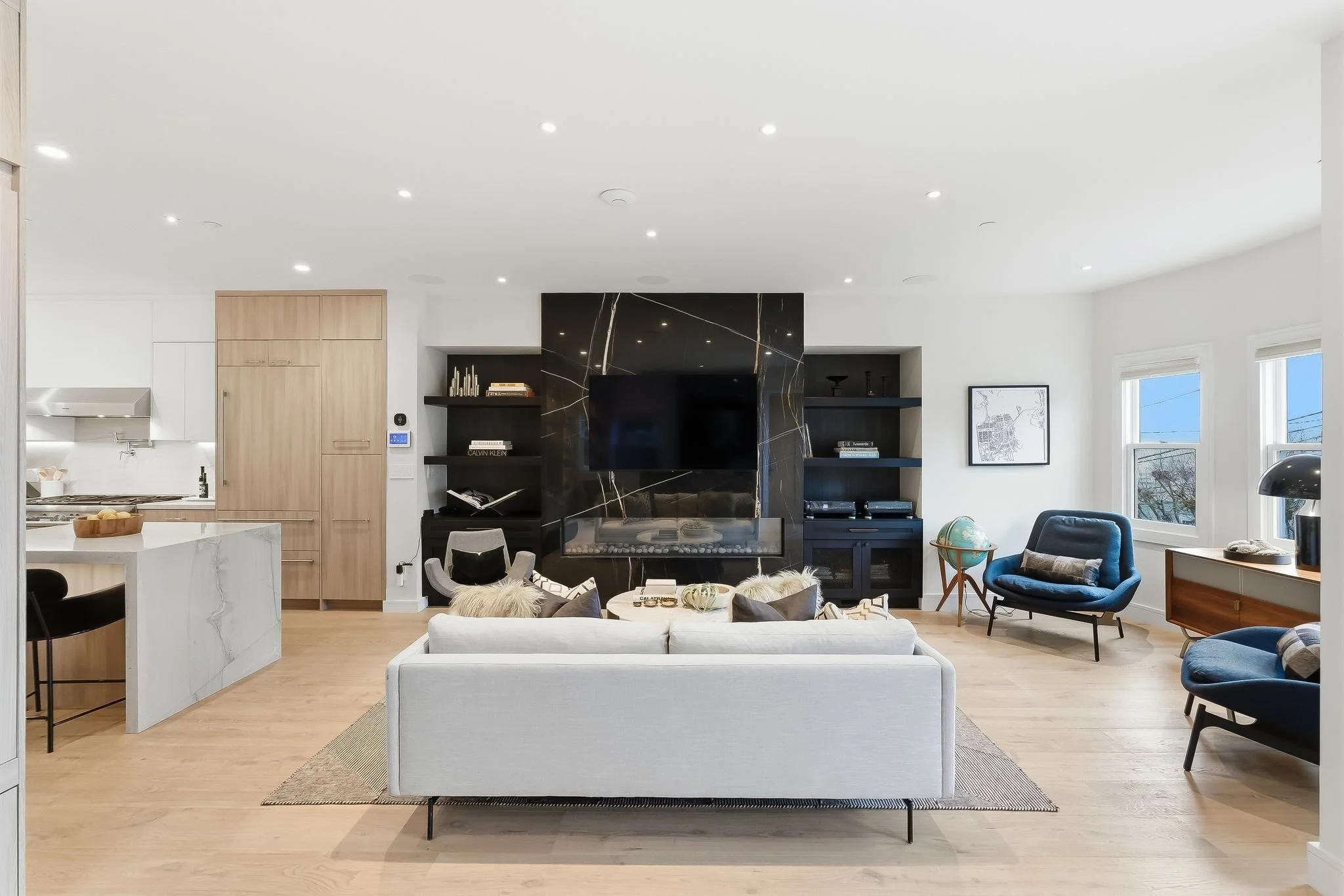 Modern living room with white sofa, black armchairs, a large black marble fireplace with TV, wooden and black shelving, and large windows letting in natural light.